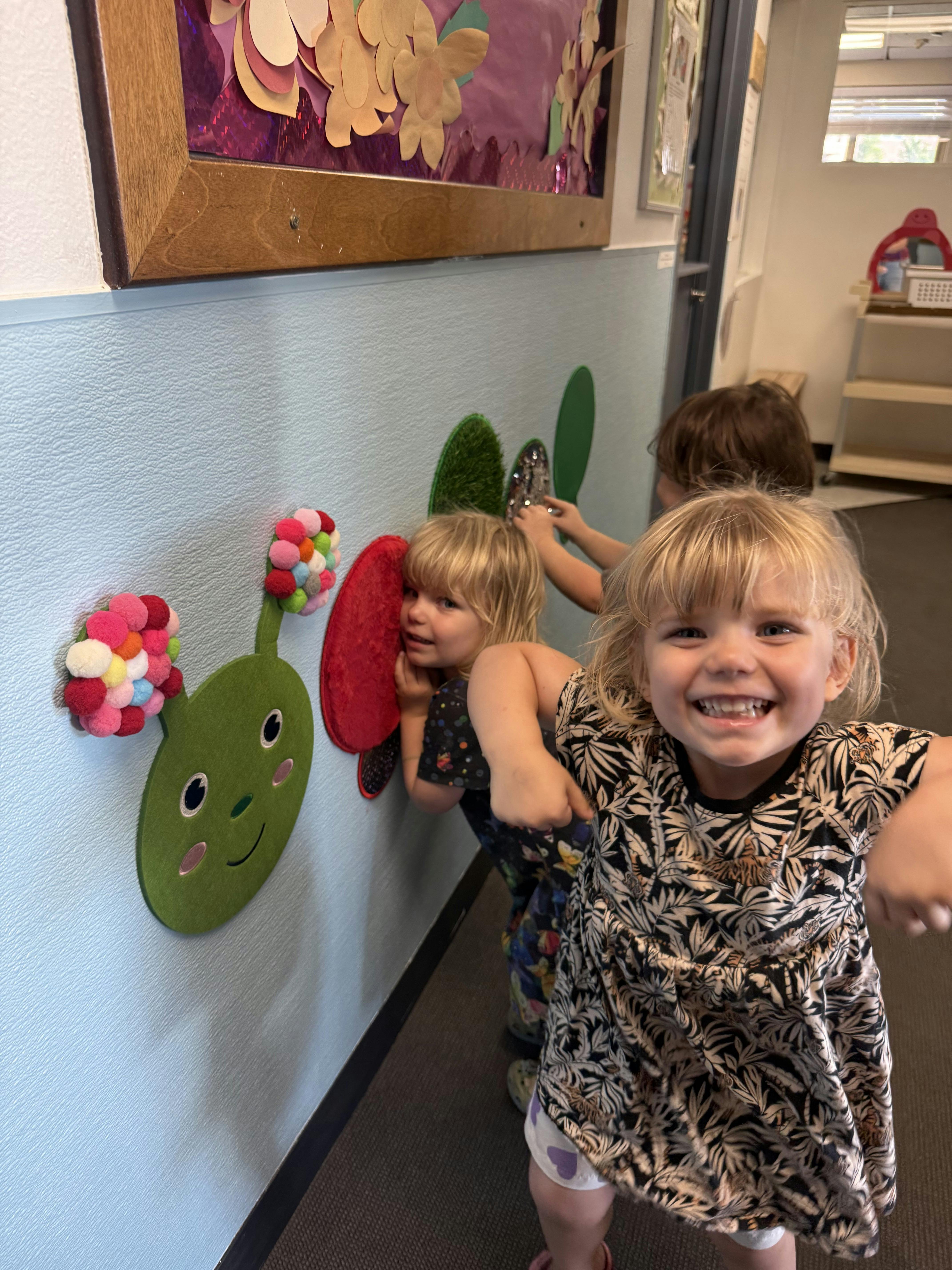 Child in patterned dress smiling at camera, other children playing with wall decor