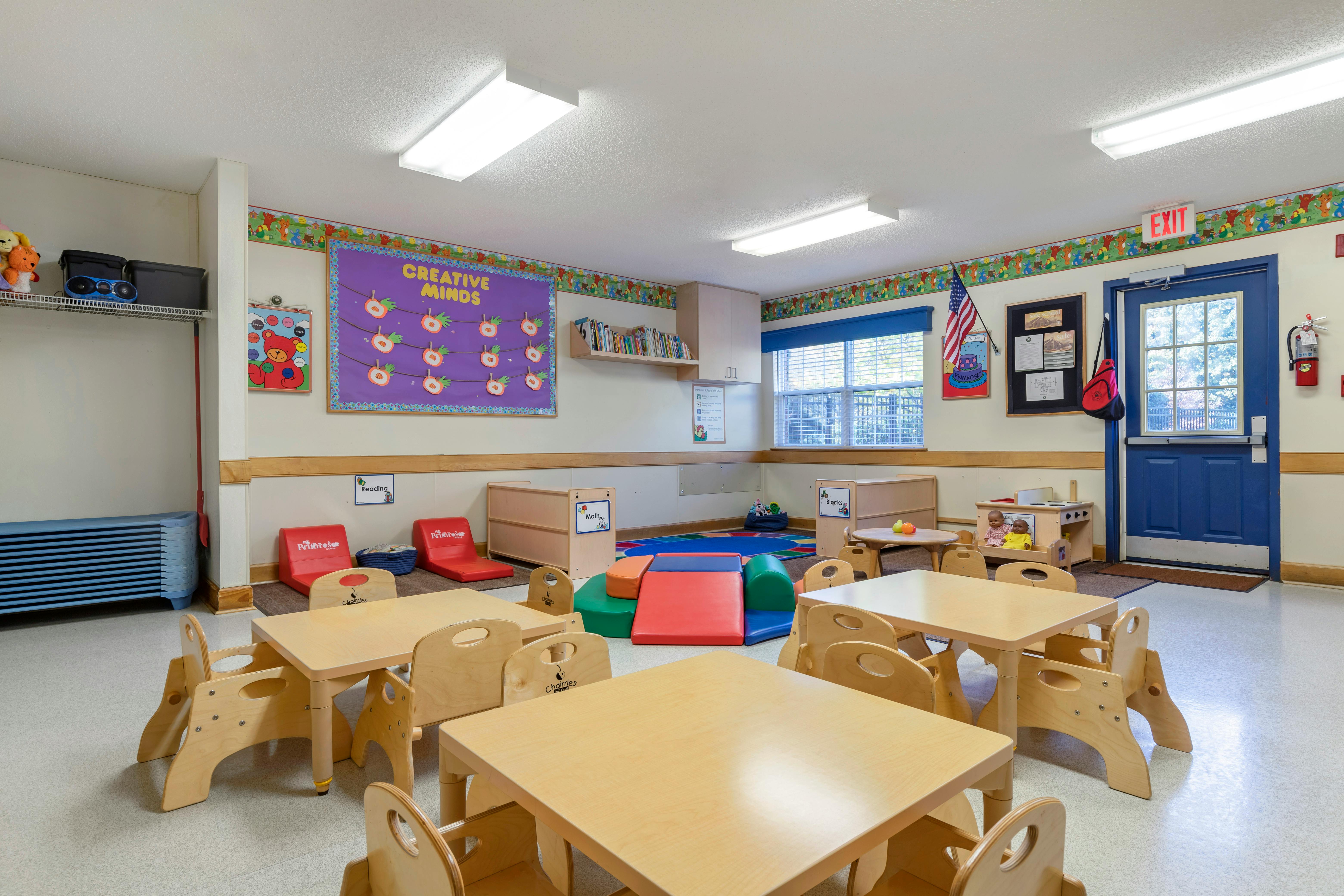 An empty early childhood classroom with small tables and chairs, play mats, and learning displays