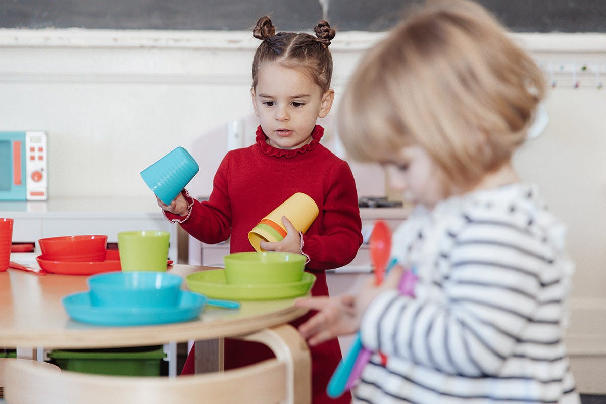 Child holding cups, another child playing with toys in a classroom