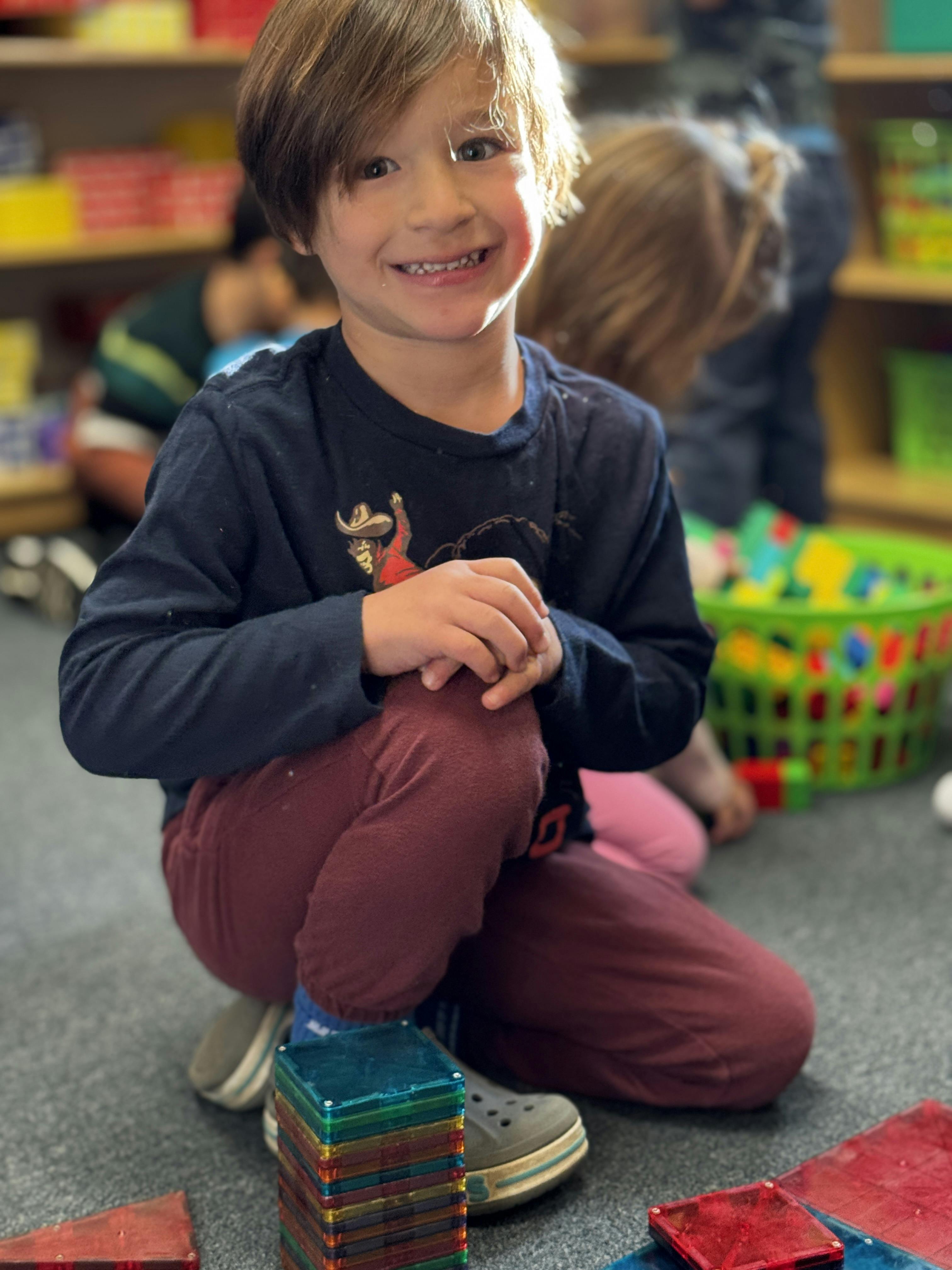 Child kneeling on a classroom floor next to colorful magnetic tiles