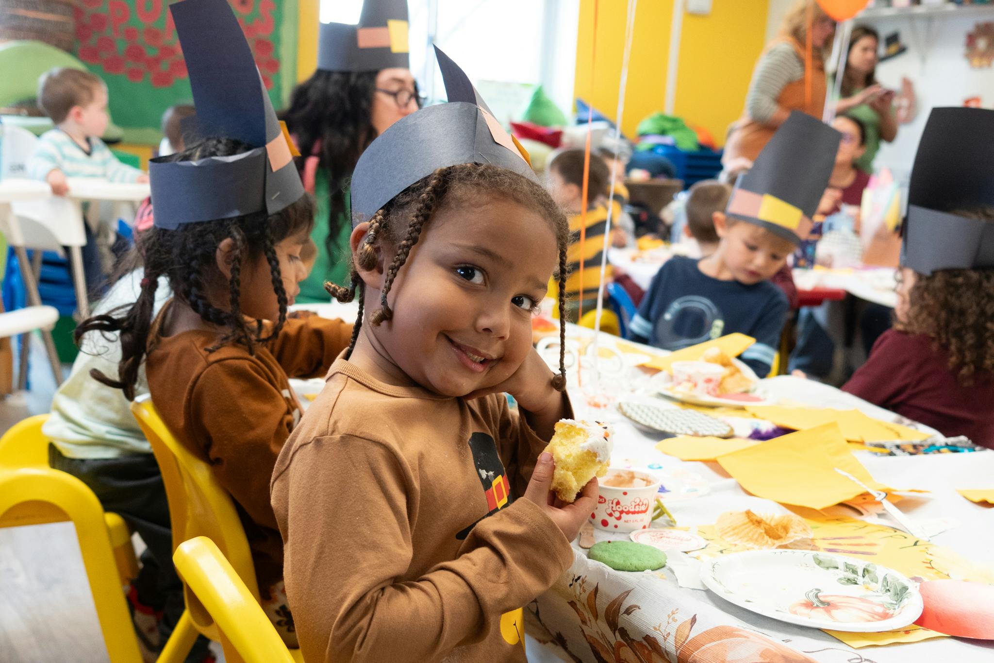 Child wearing a paper hat eating cake at a table in a classroom