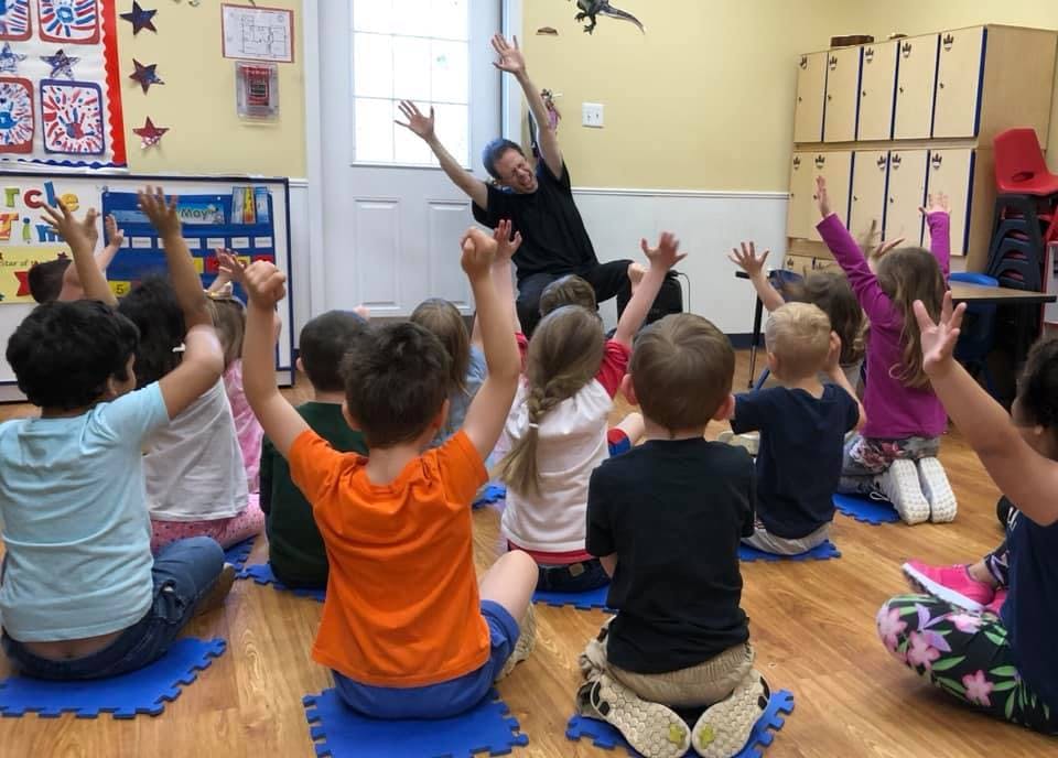 Adult and children sitting on floor with arms raised in a classroom