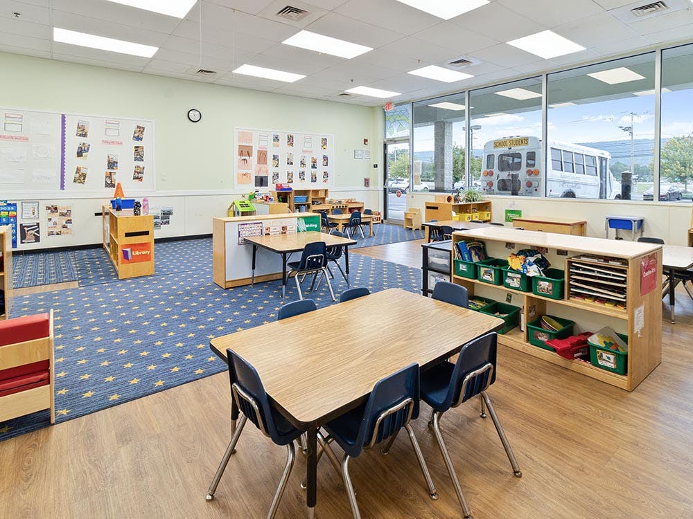 Classroom with small tables and chairs, shelves with books and bins, large windows