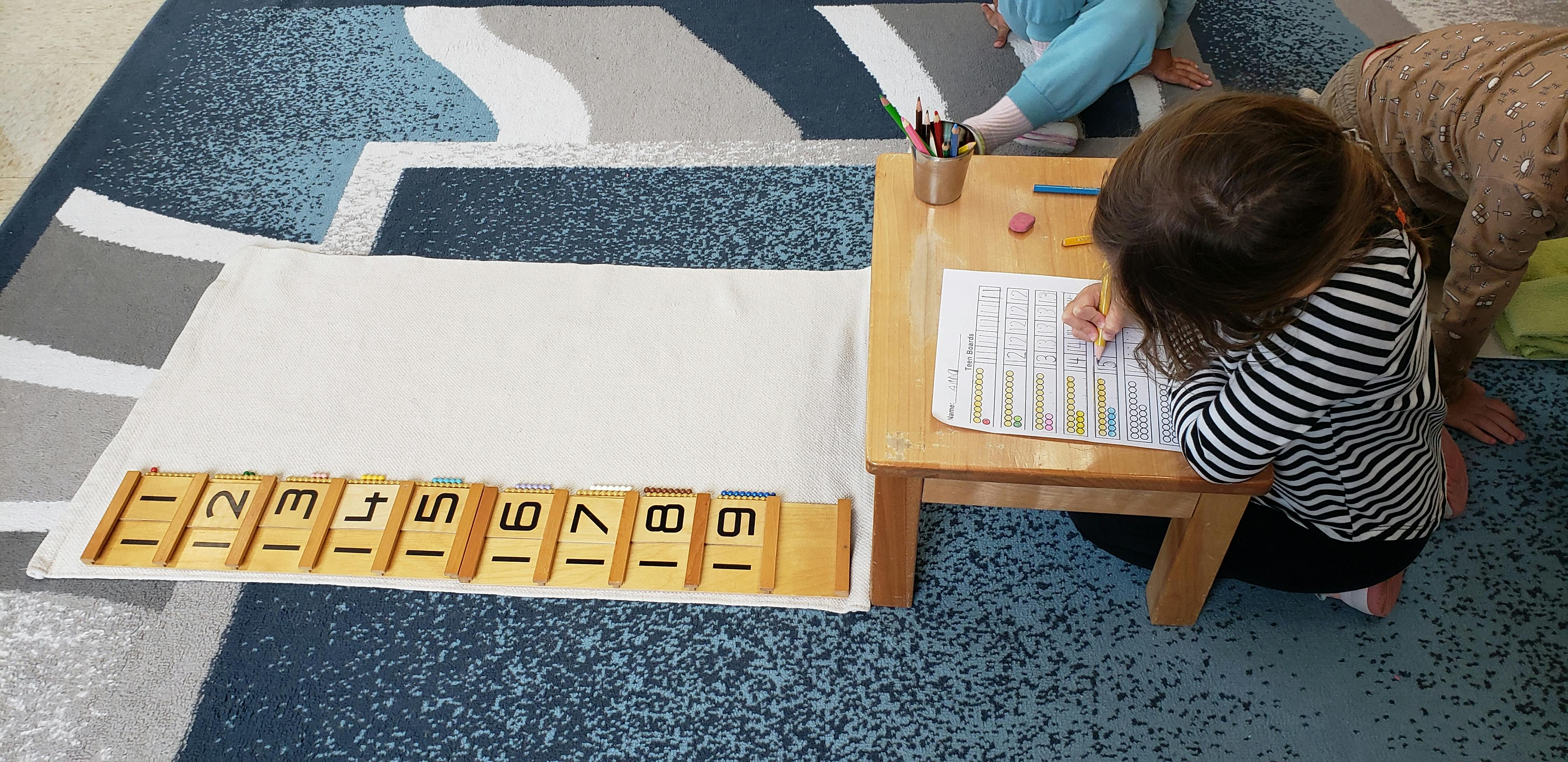 Child sitting at a small table writing on a worksheet with numbered tiles