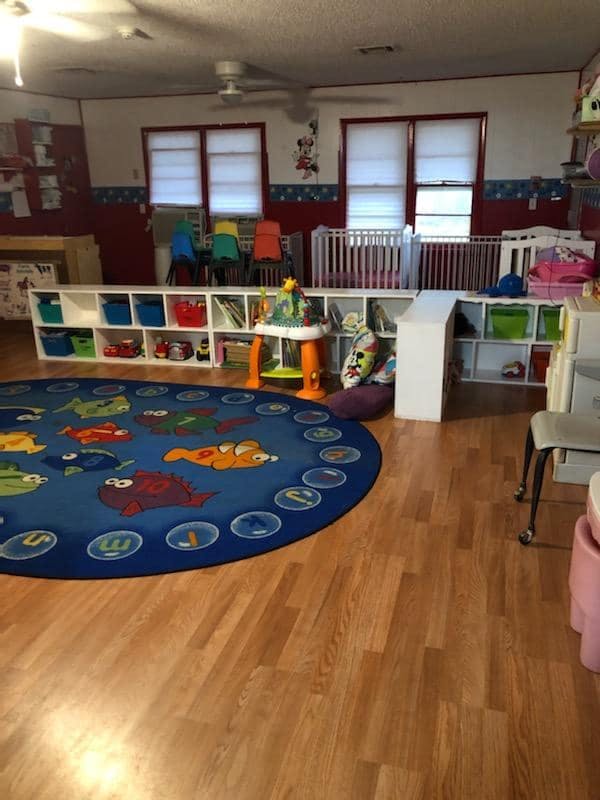 Daycare room with cribs, shelves of toys, and a colorful rug