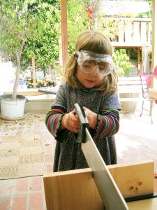 Child wearing safety goggles using a saw to cut wood