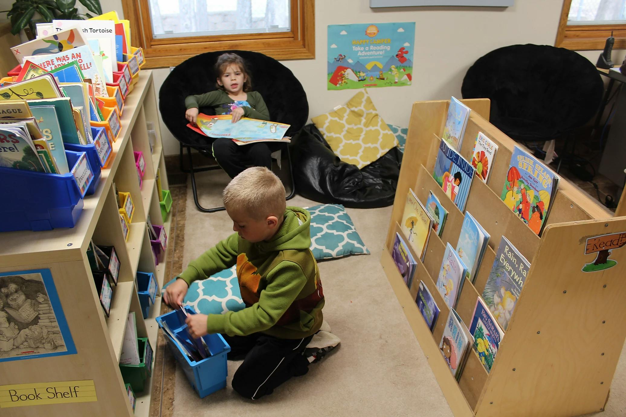 Child kneeling and looking into a blue bin, another child reading in a chair
