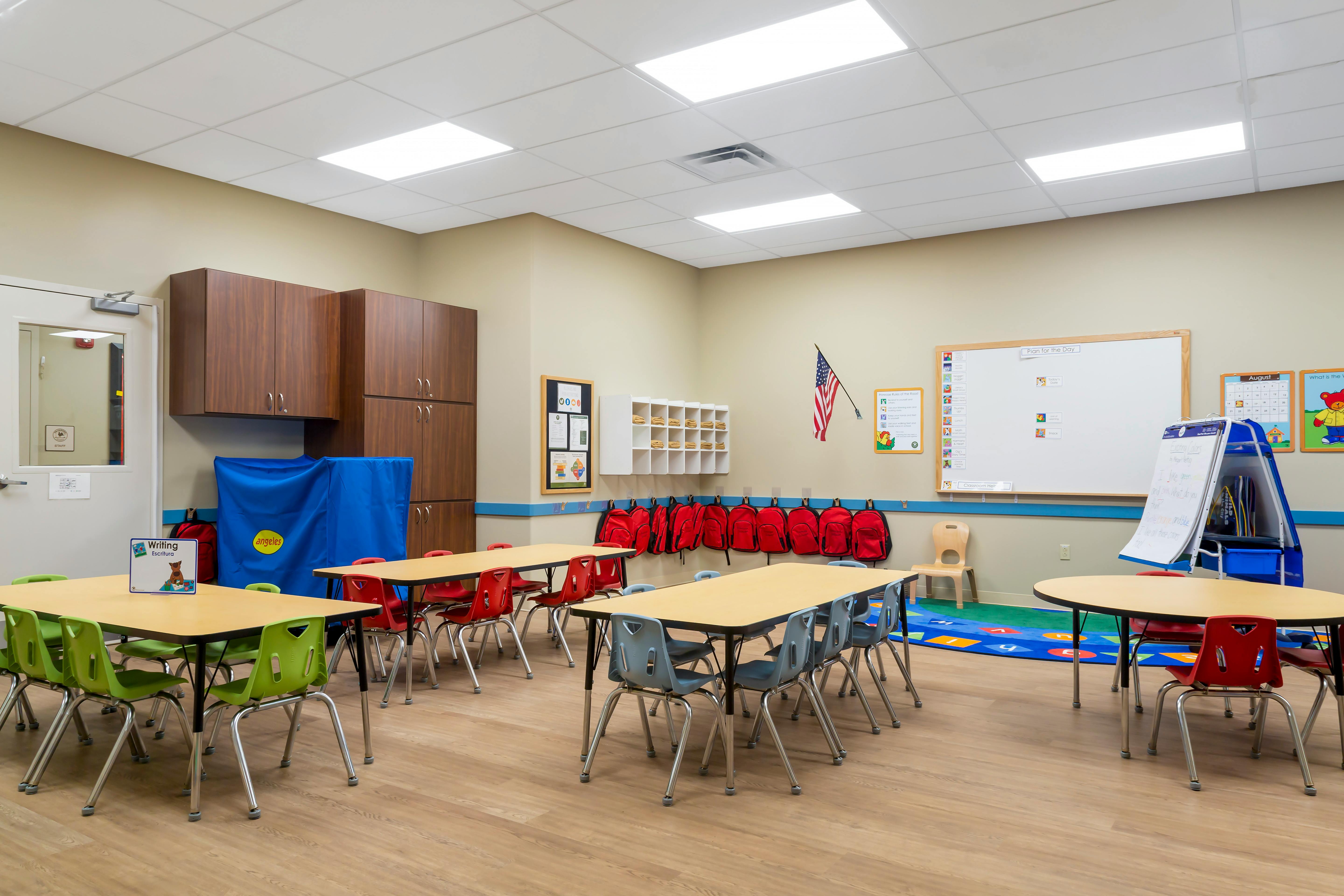Classroom with tables and chairs, red backpacks hanging on a wall