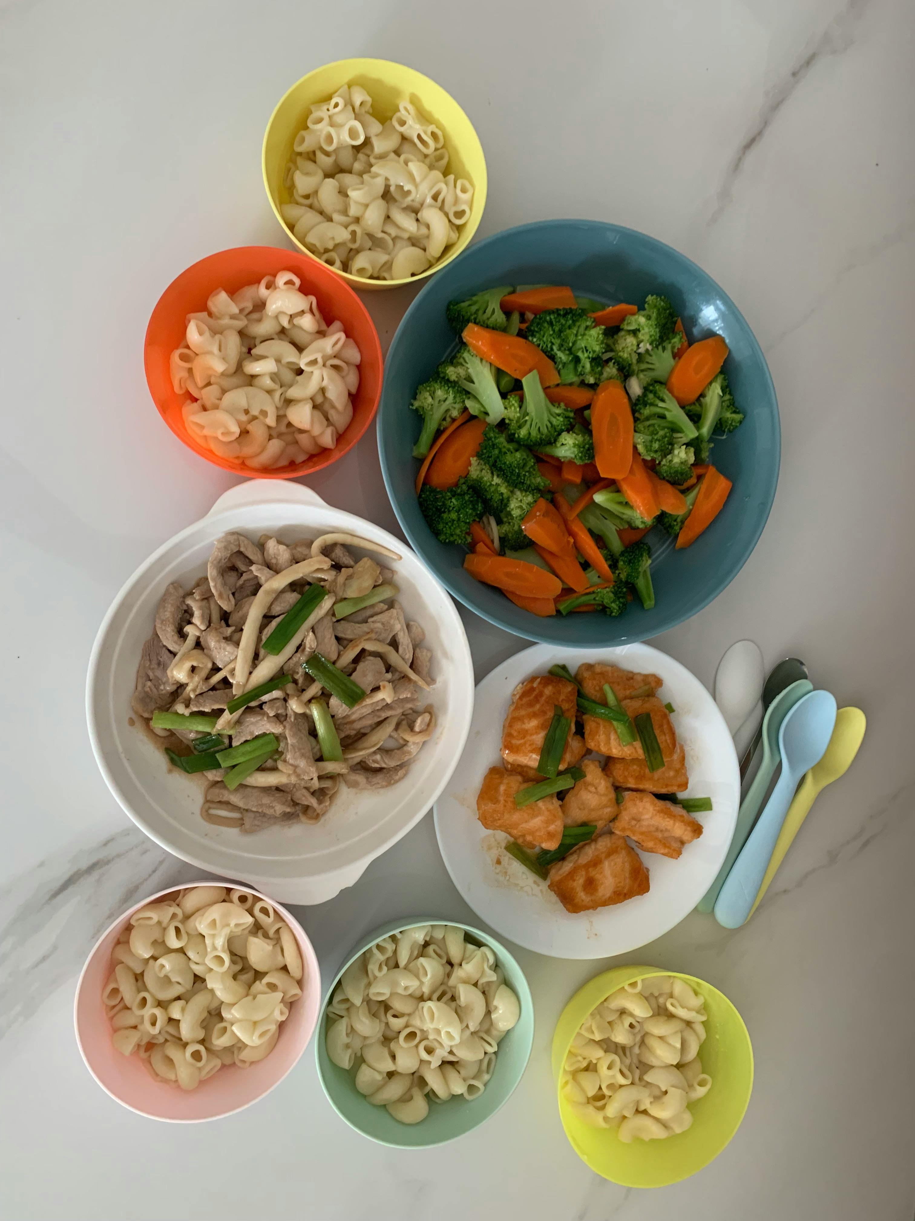 Several bowls of macaroni and plates of cooked broccoli, carrots, meat, and fish