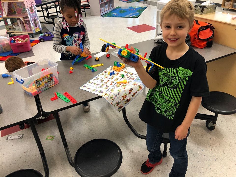 Child holding a toy airplane while another child plays with toys at a table