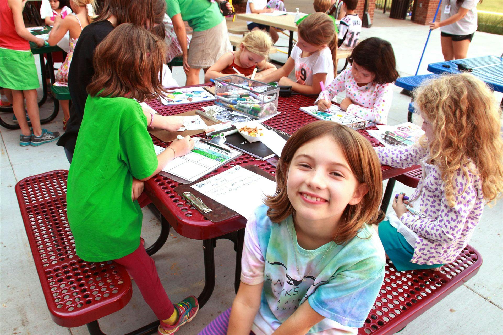 Child smiling at camera while other children draw at a table