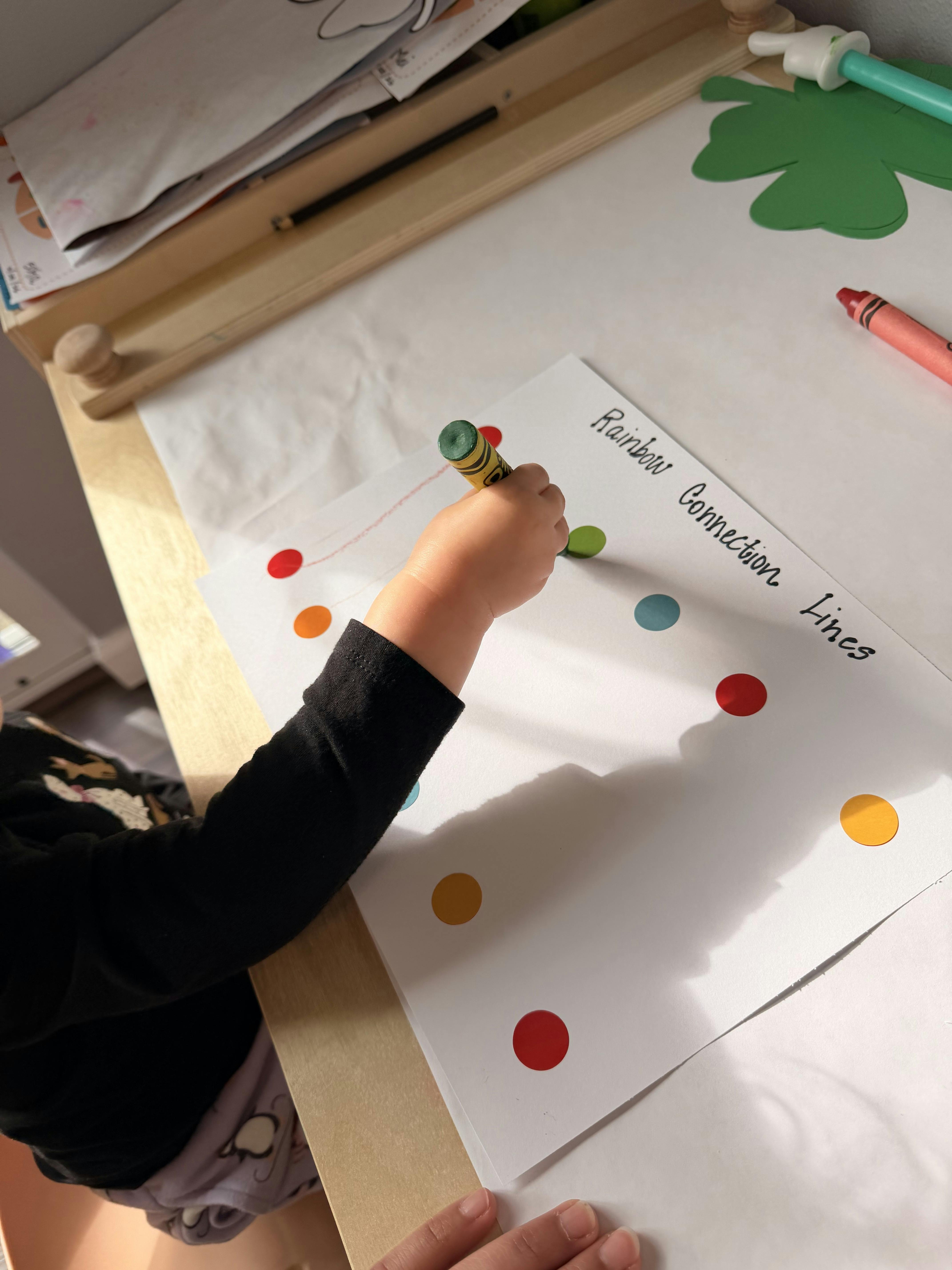 Child's hand holding a green crayon, drawing on paper with colorful dots