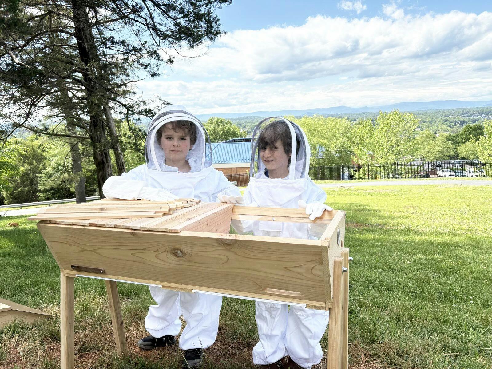 Two children in beekeeper suits standing by a beehive