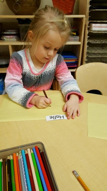 Child writing on paper with a pencil, name tag "Halle" on the table