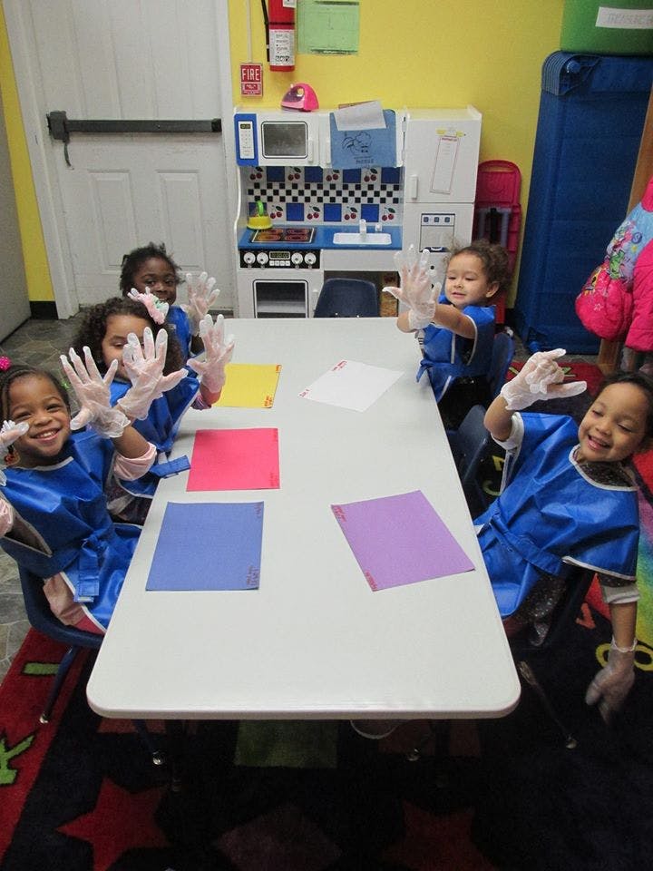Children in blue smocks and gloves raising hands at a table