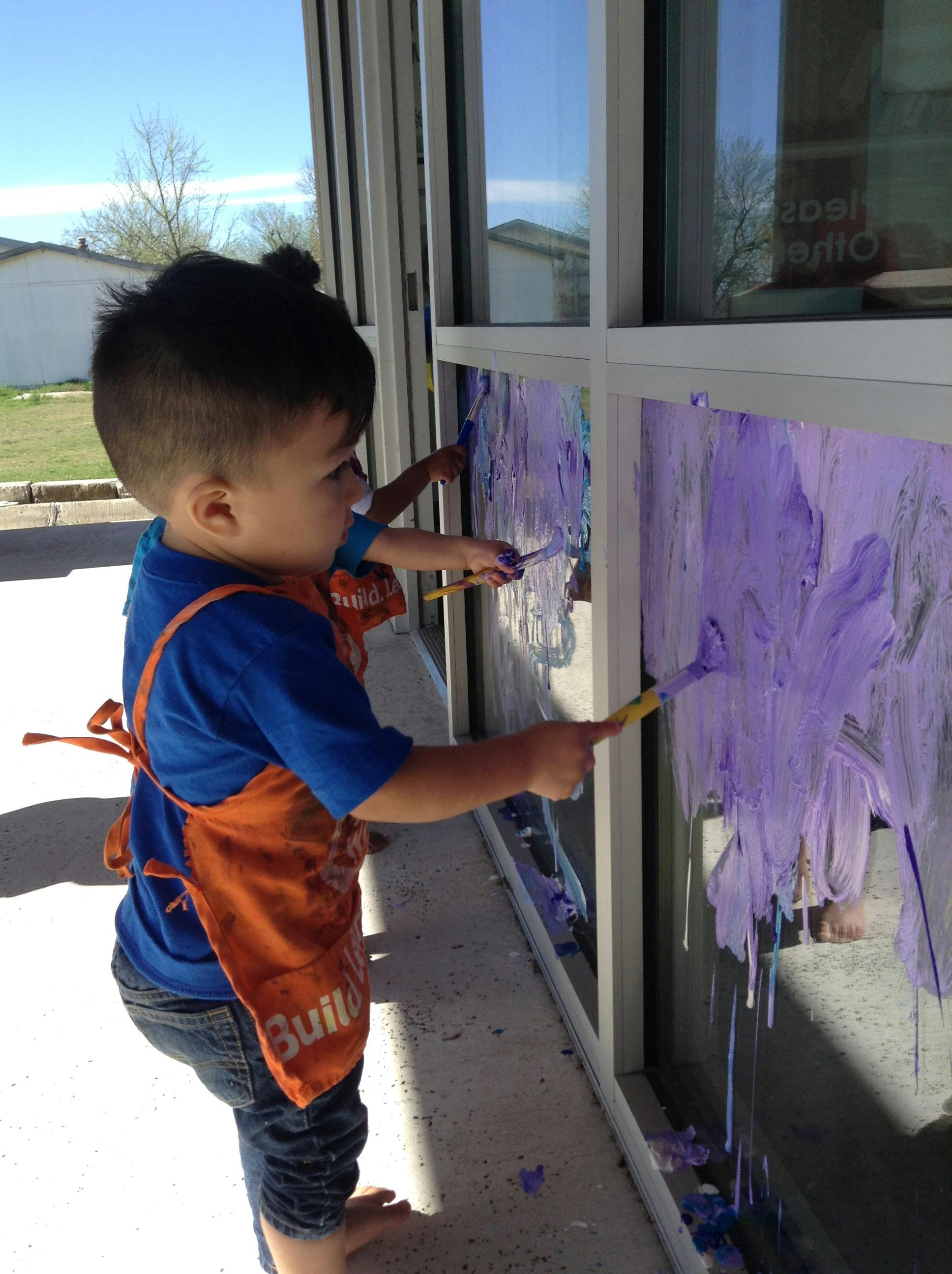 Child in an apron painting on a window with purple paint