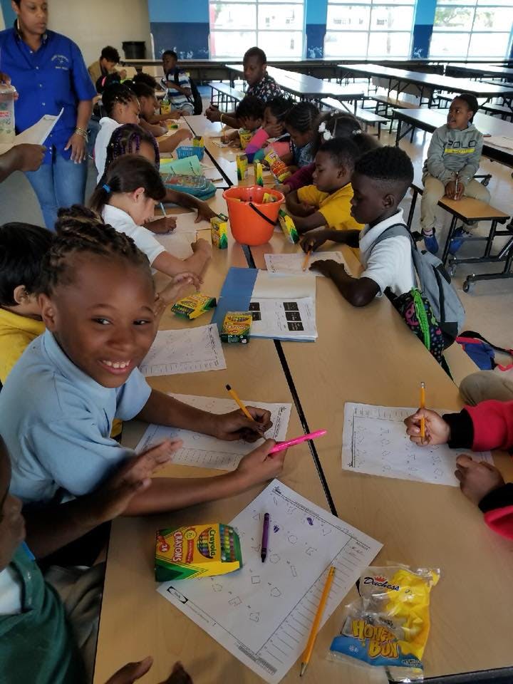Children writing at tables in a classroom with an adult supervising