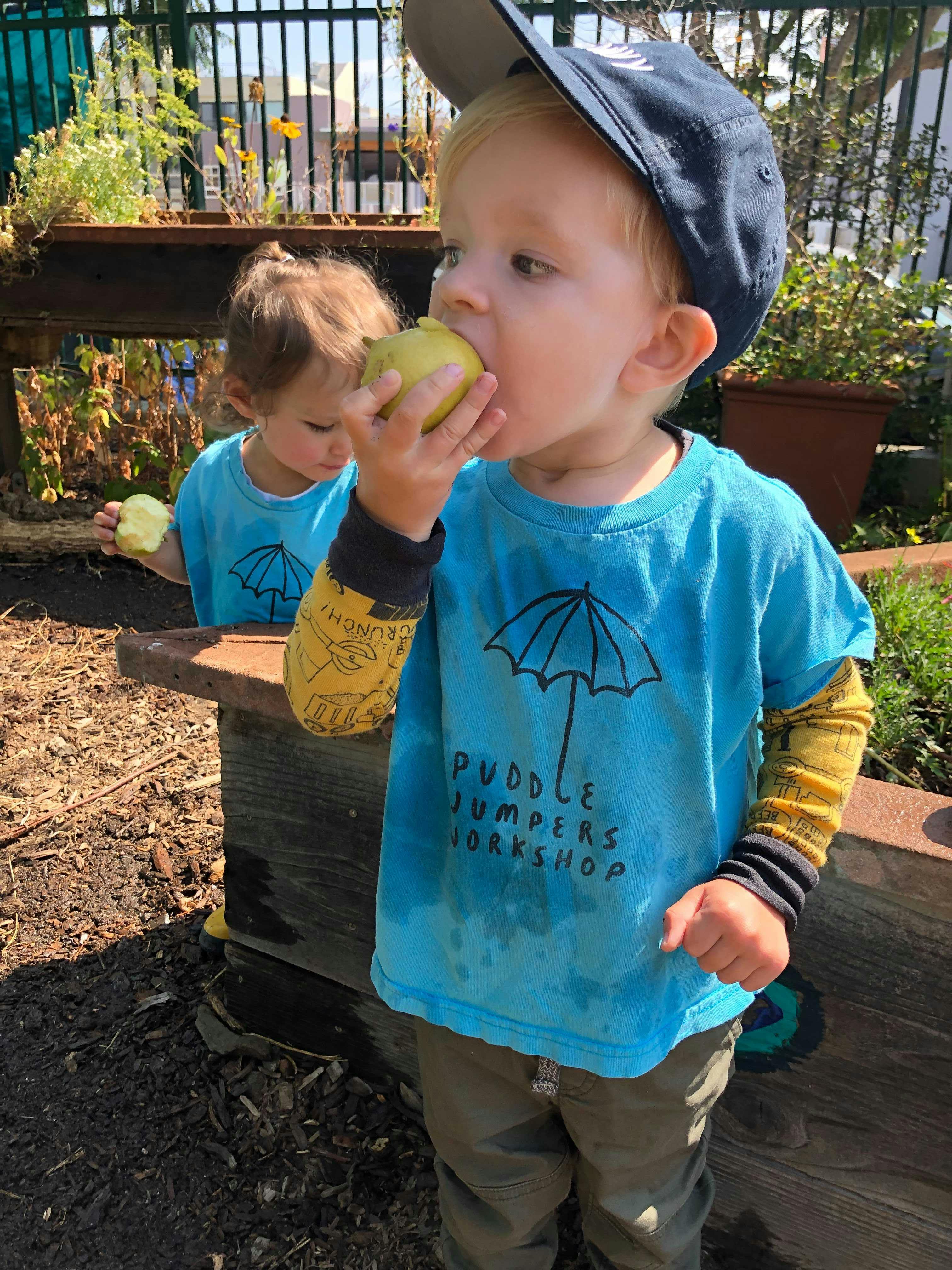 Child in a blue shirt and hat eating an apple, another child in background