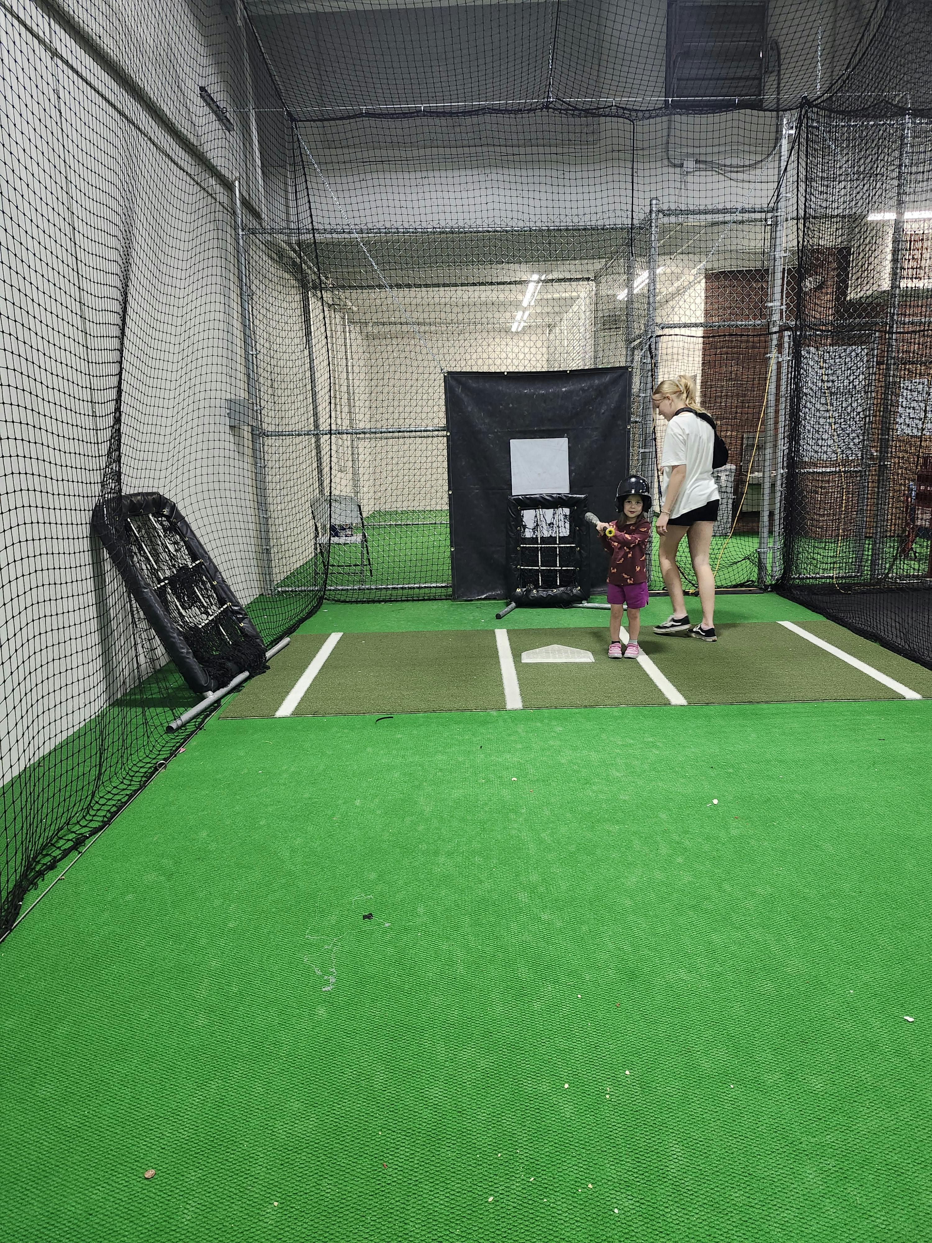 A person and a child with a helmet and bat in a batting cage