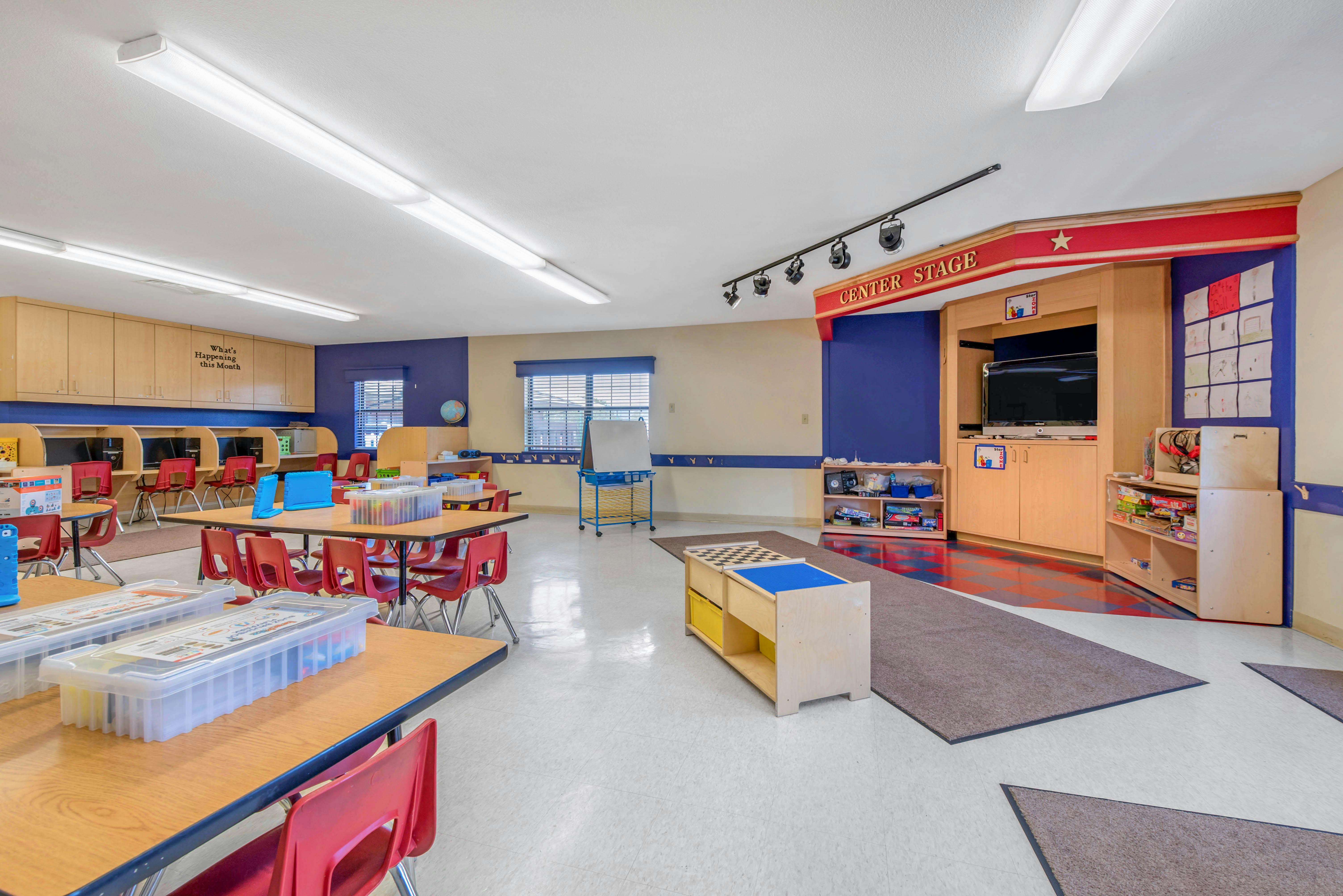 Classroom with small tables and chairs, computer stations, and a stage area