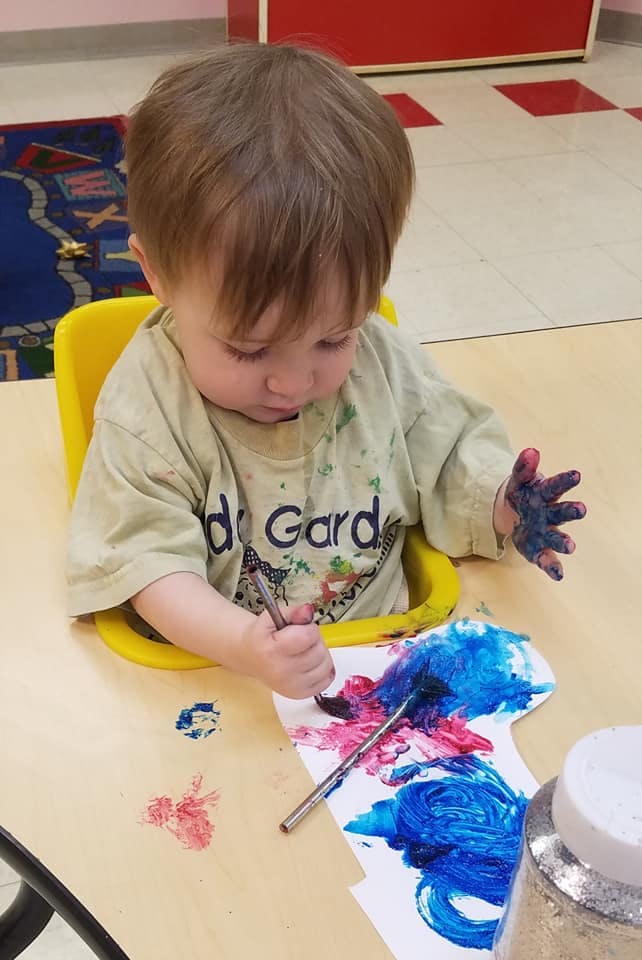 Child in a high chair painting with a brush and blue paint on hands