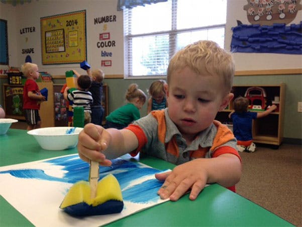 Child painting with a sponge brush at a table in a classroom