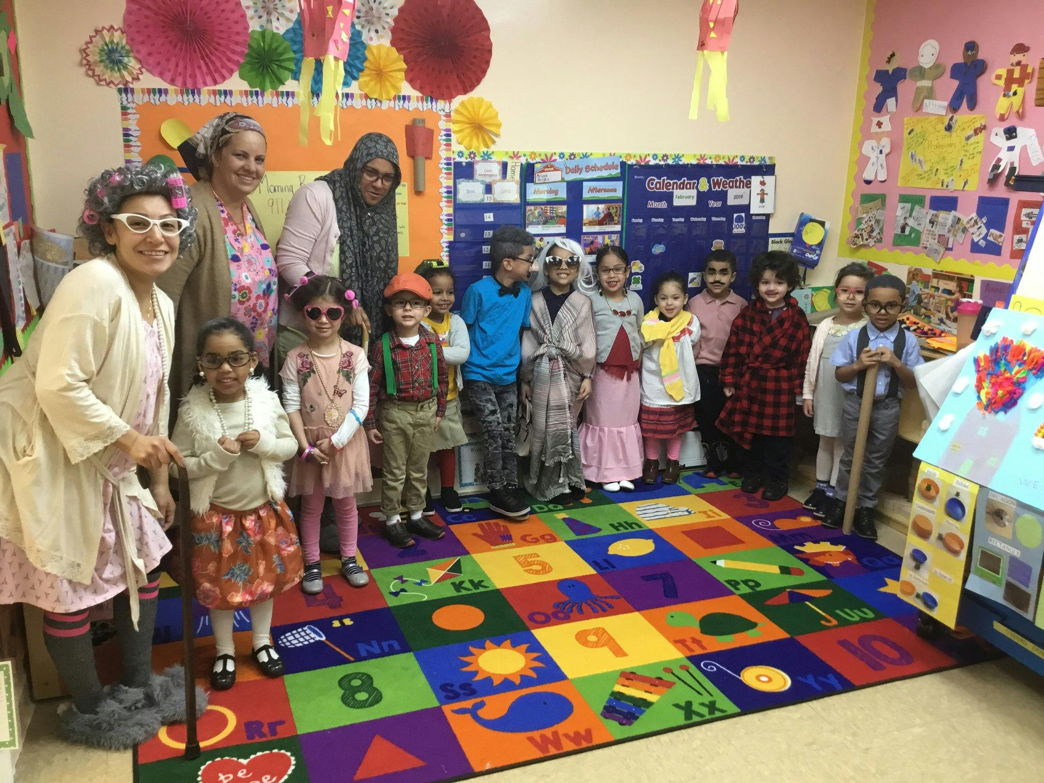 Adults and children in costumes standing on a colorful rug in a classroom