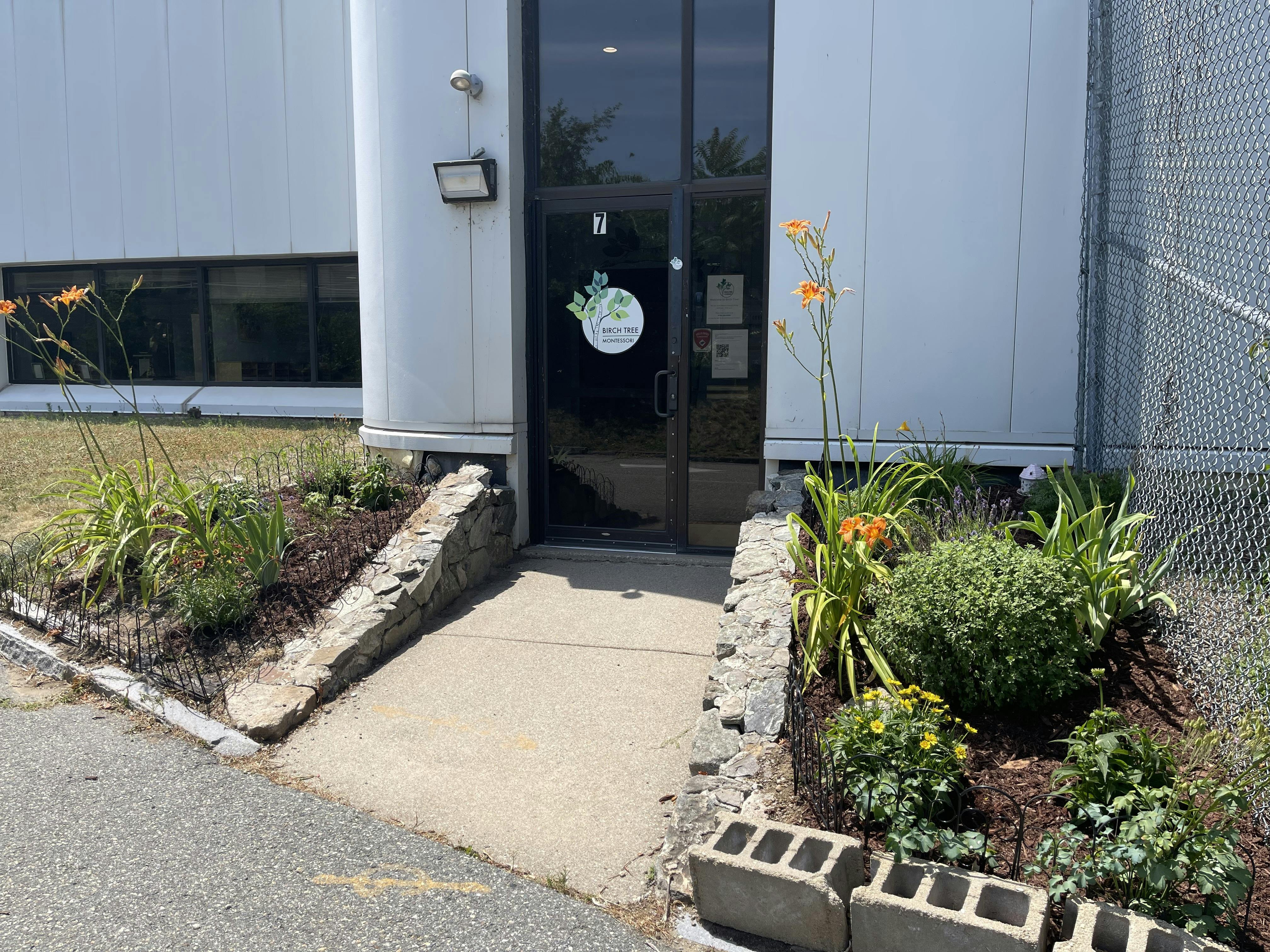 Building entrance with a glass door and Birch Tree Montessori logo, surrounded by garden beds