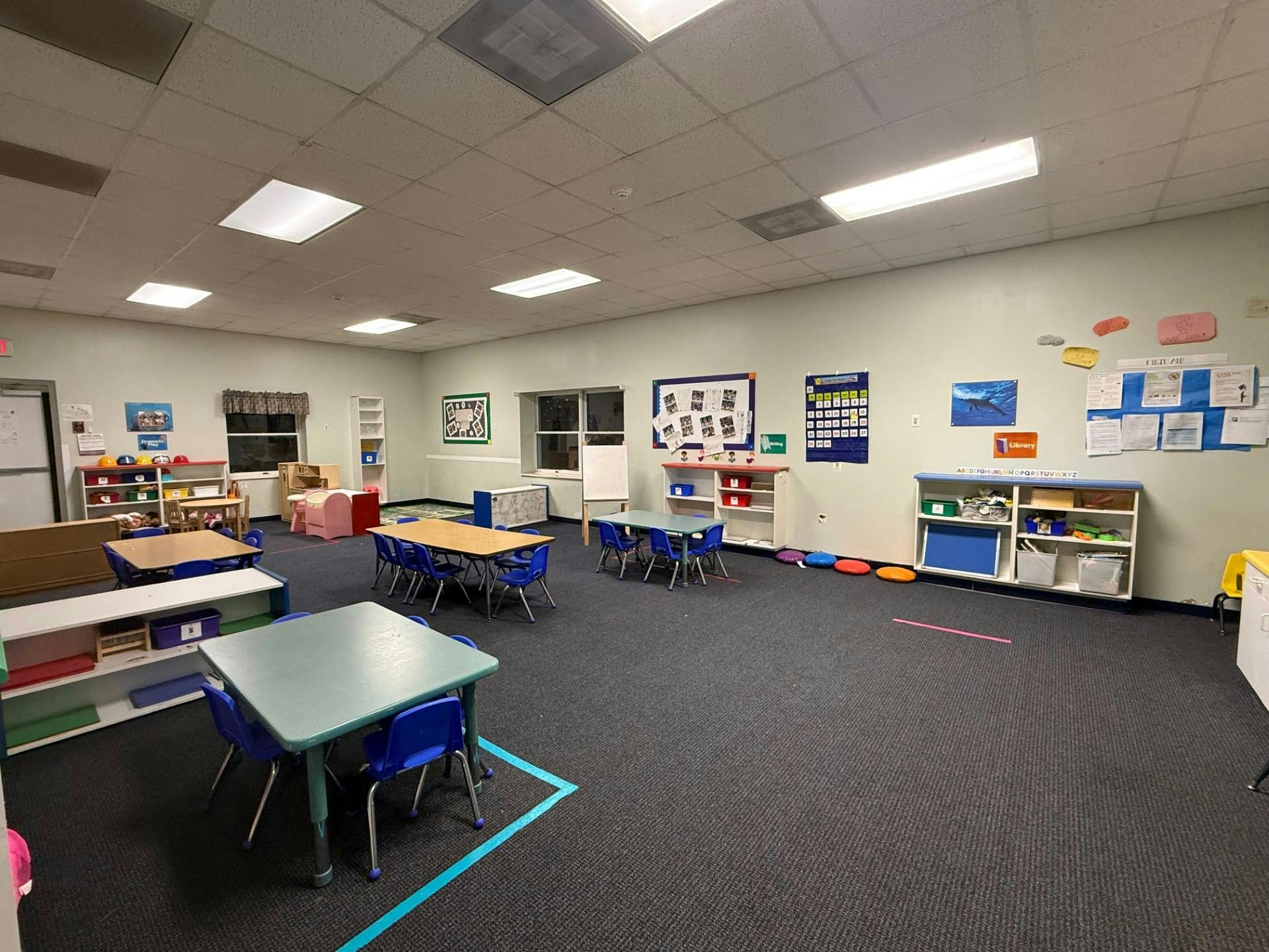 Empty classroom with small tables and chairs, shelves with toys, and posters on the wall