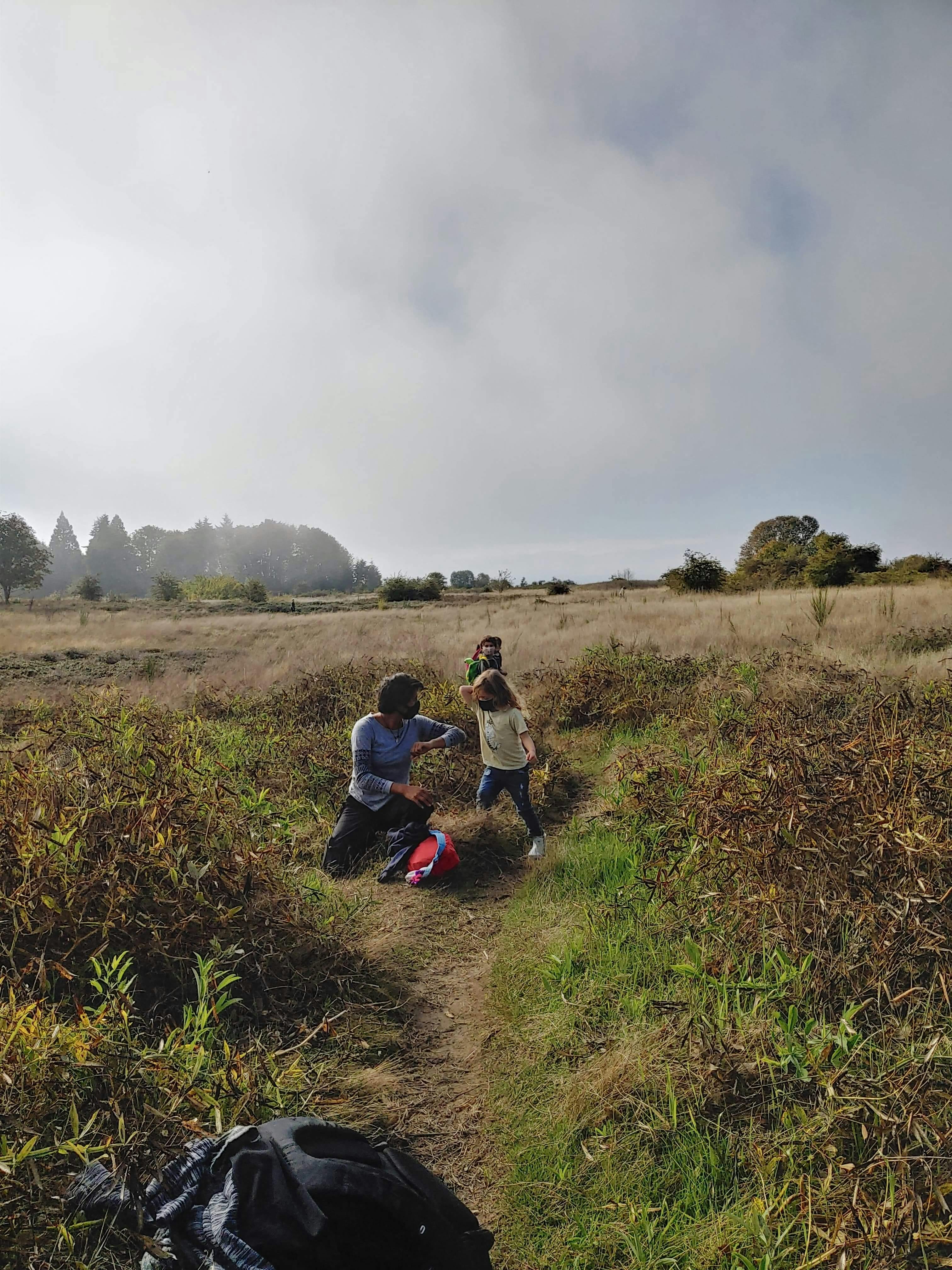 Person kneeling on a dirt path with two children in a grassy field
