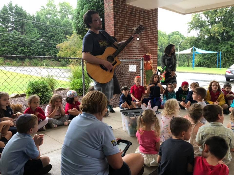 Person playing guitar for a group of children sitting on the ground outdoors