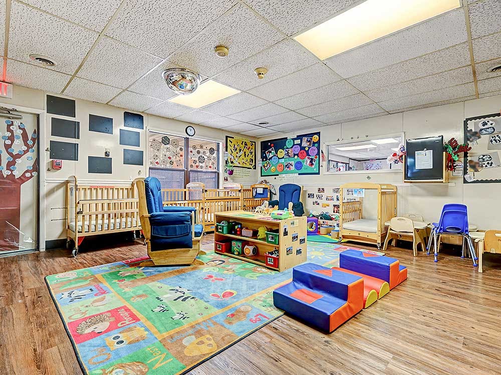 A colorful childcare classroom with cribs, play rug, and child-sized furniture
