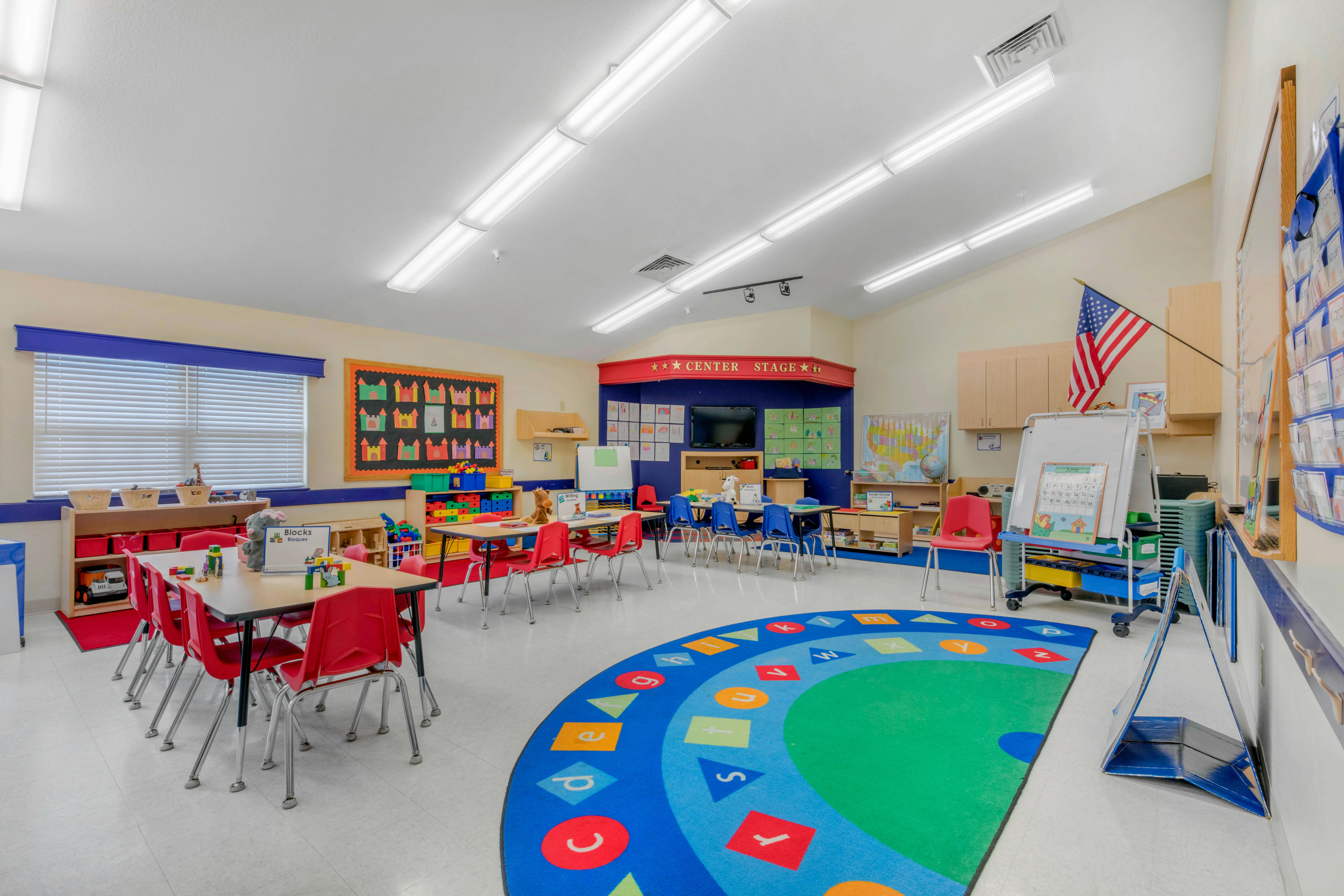 A colorful kindergarten classroom with small tables and chairs, and a round ABC rug