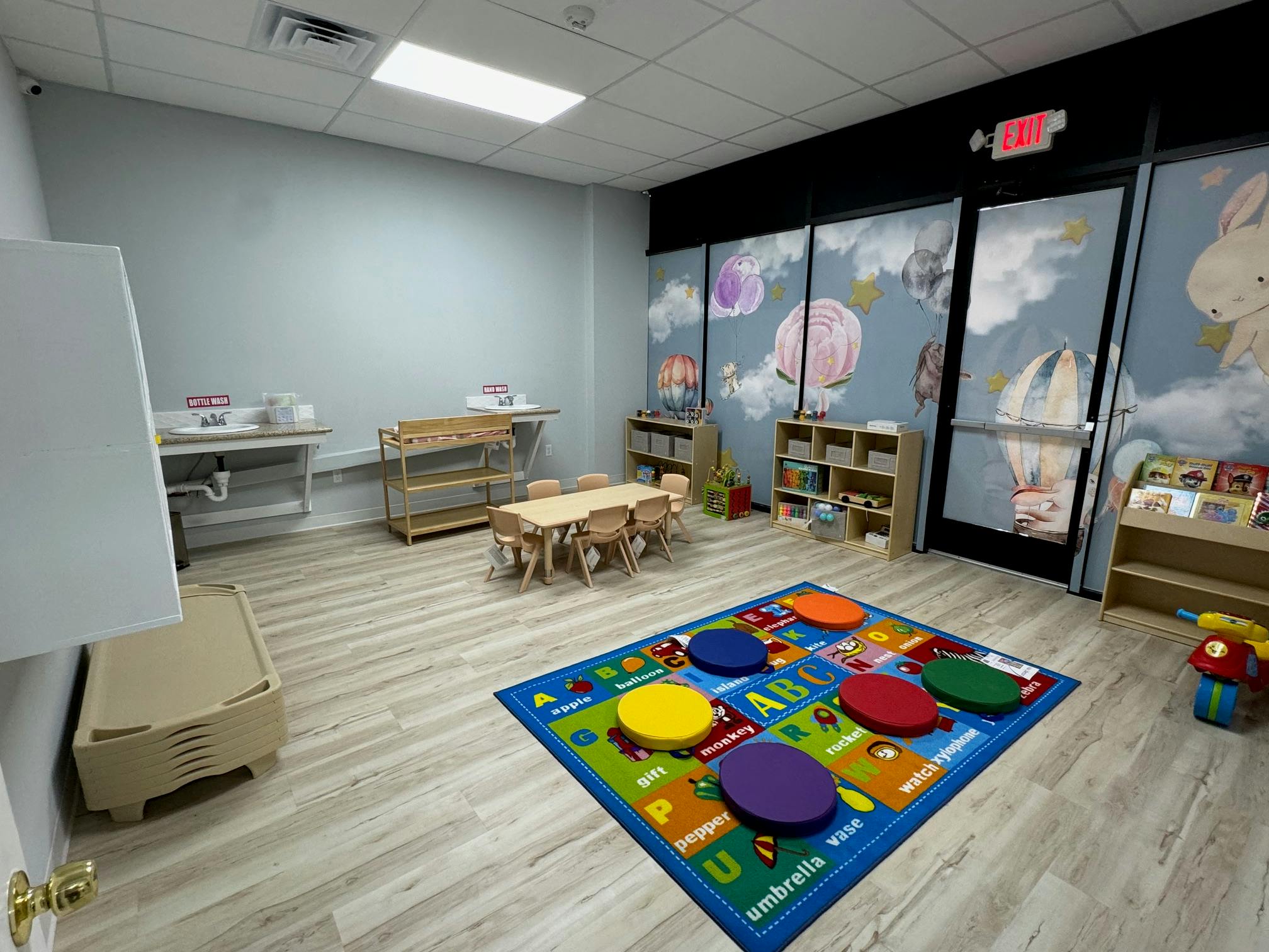 Empty classroom with small tables and chairs, a colorful rug, and shelves