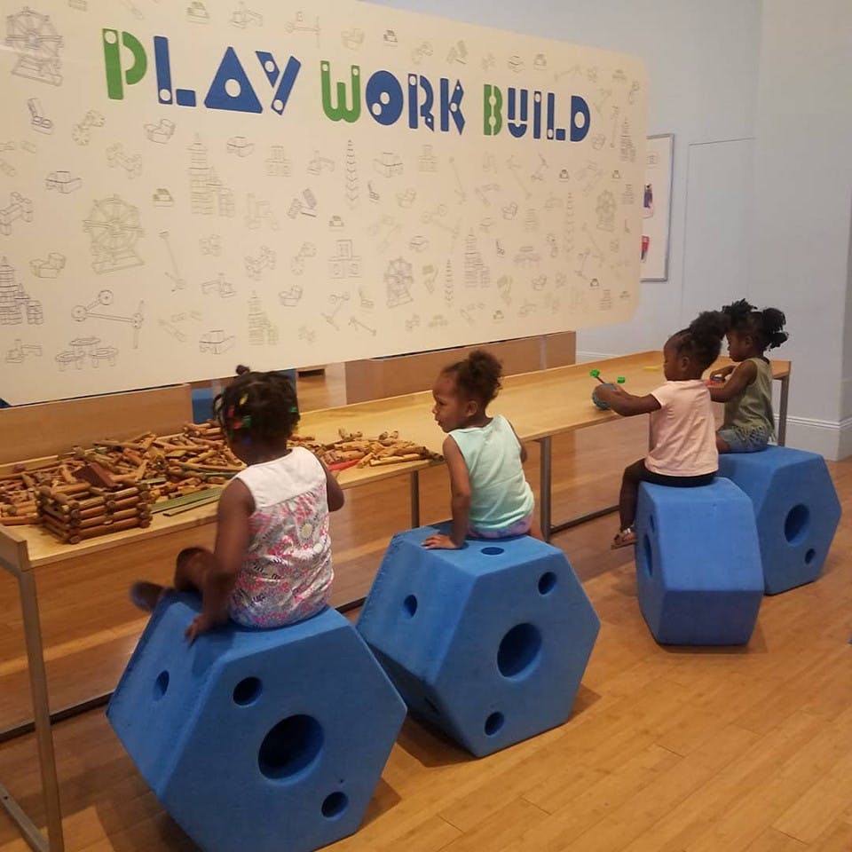 Children sitting on blue hexagonal blocks in front of a wall with "PLAY WORK BUILD"