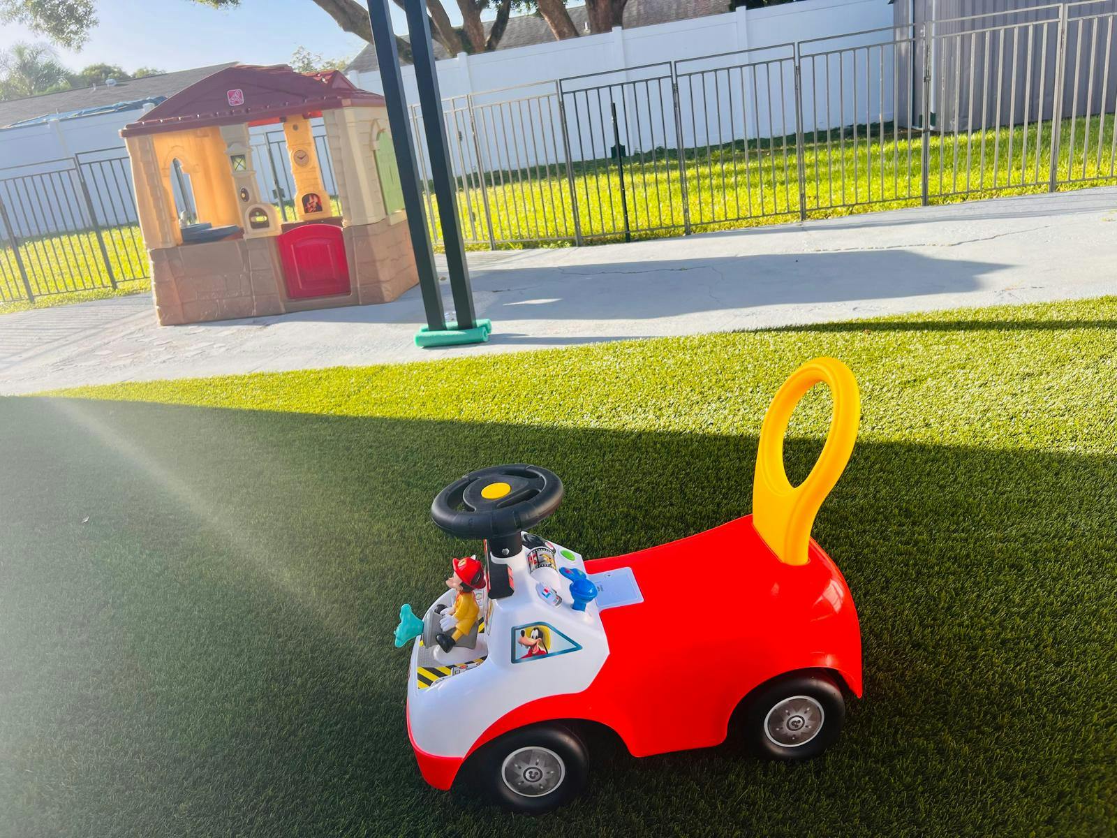 Red and white ride-on toy car on artificial grass with a playhouse in the background