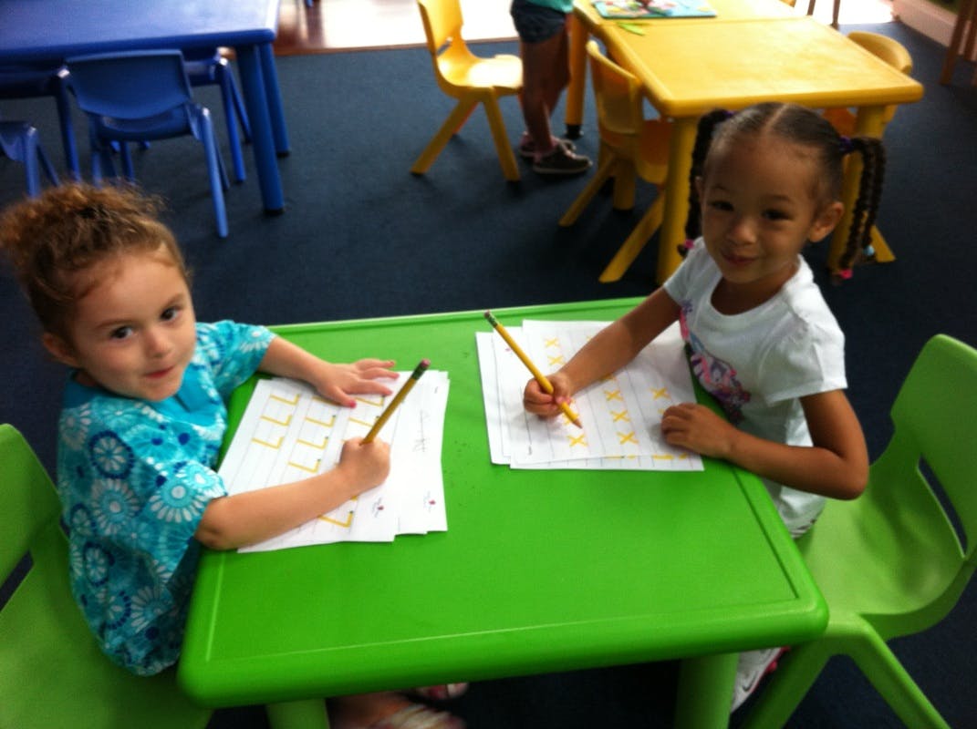 Two children writing on paper with pencils at a green table in a classroom