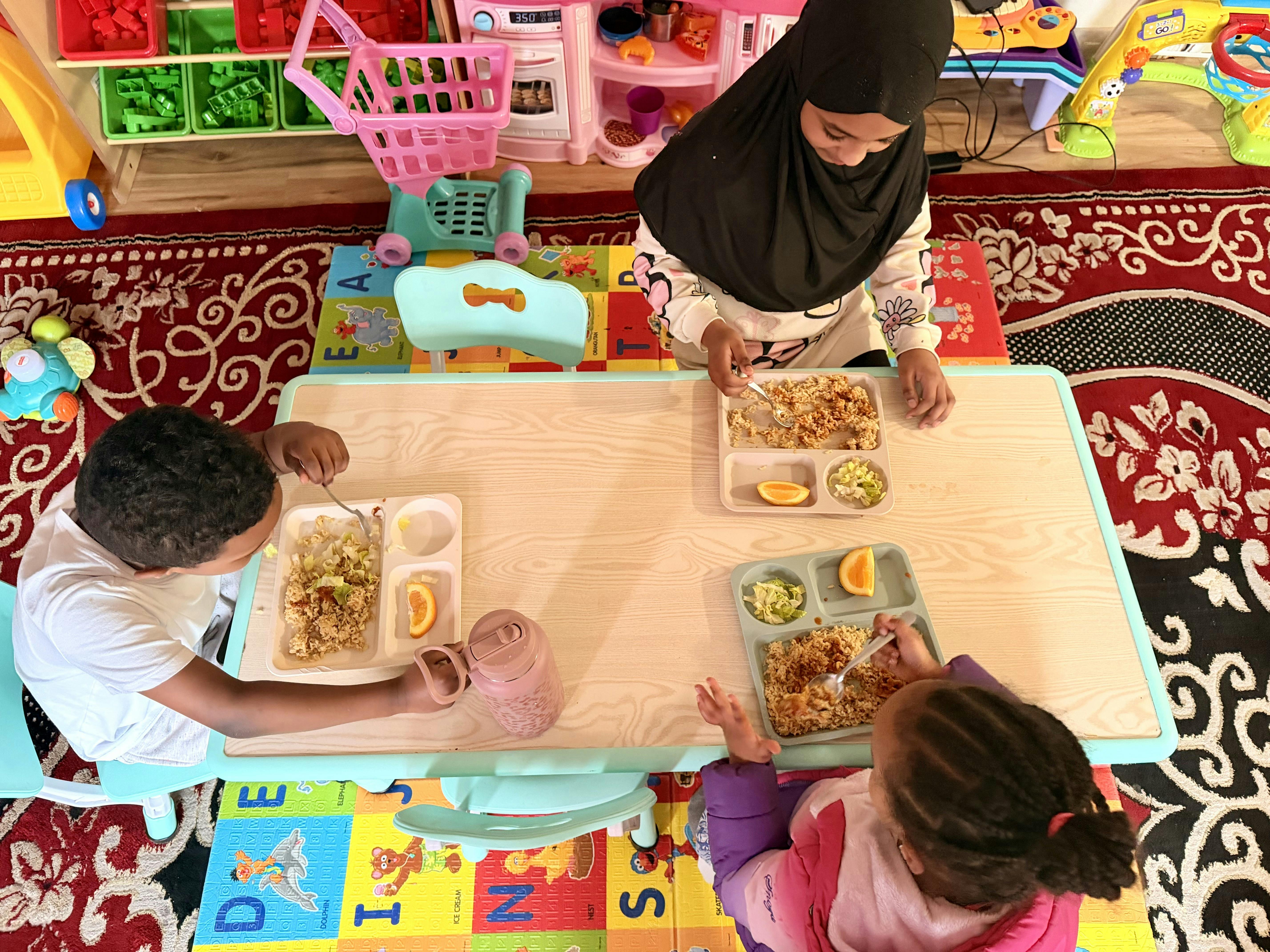 Three children eating a meal with rice, lettuce, and an orange slice at a table