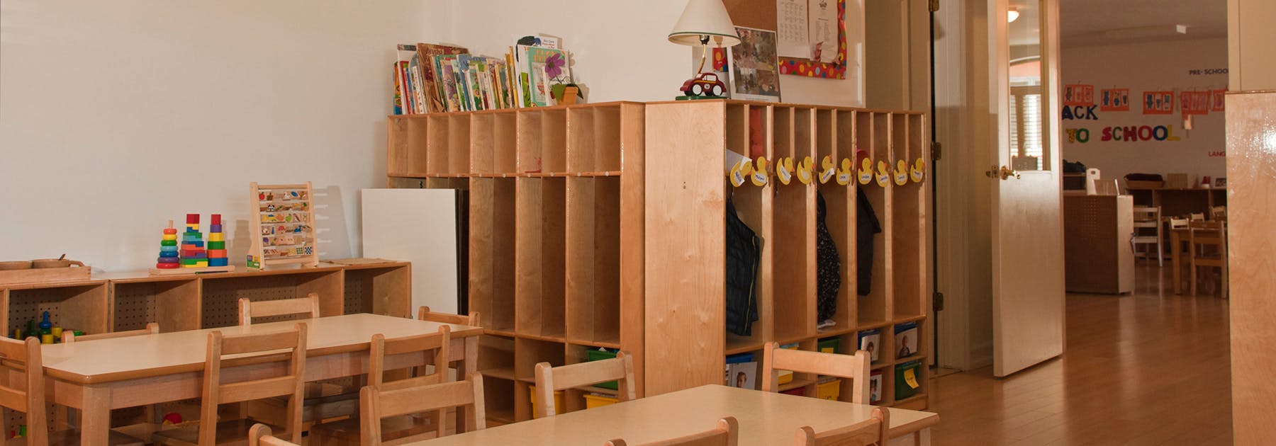 An empty classroom with small wooden tables and chairs, cubbies, and toys