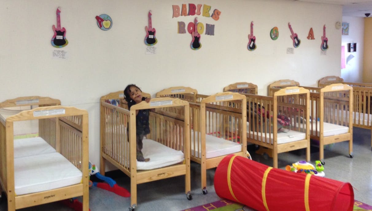 Child standing in a crib in a room with multiple cribs