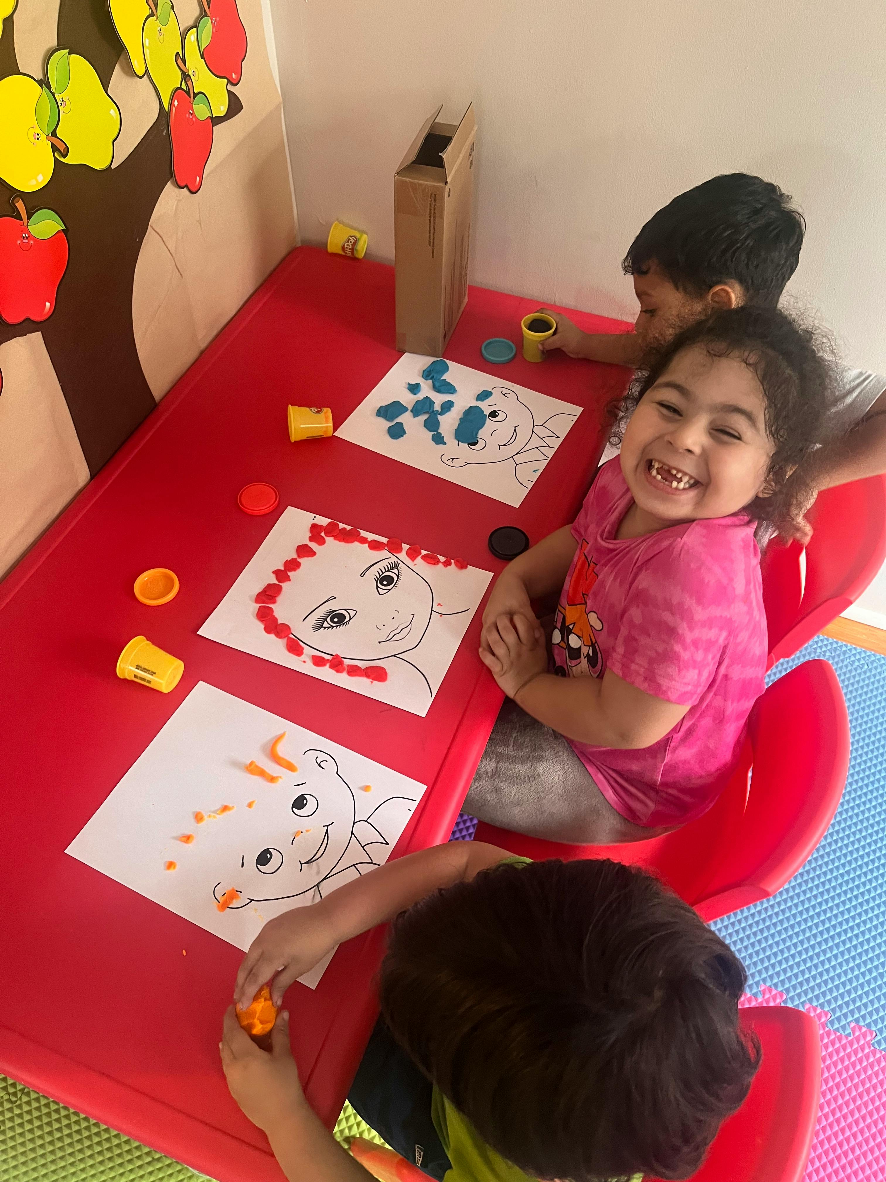 Three children at a red table doing an art activity with playdough