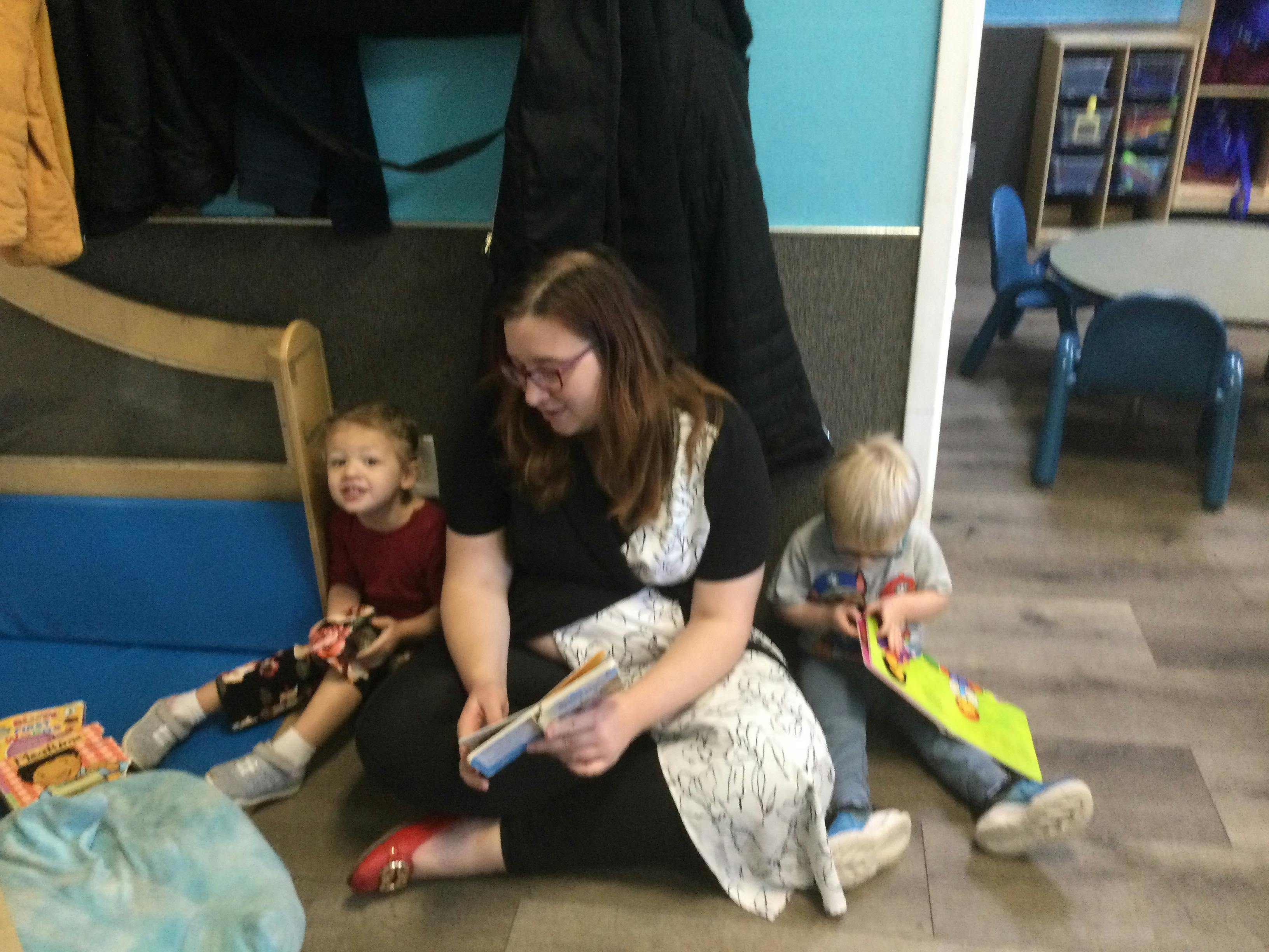 Adult and two children sitting on the floor reading books in a classroom