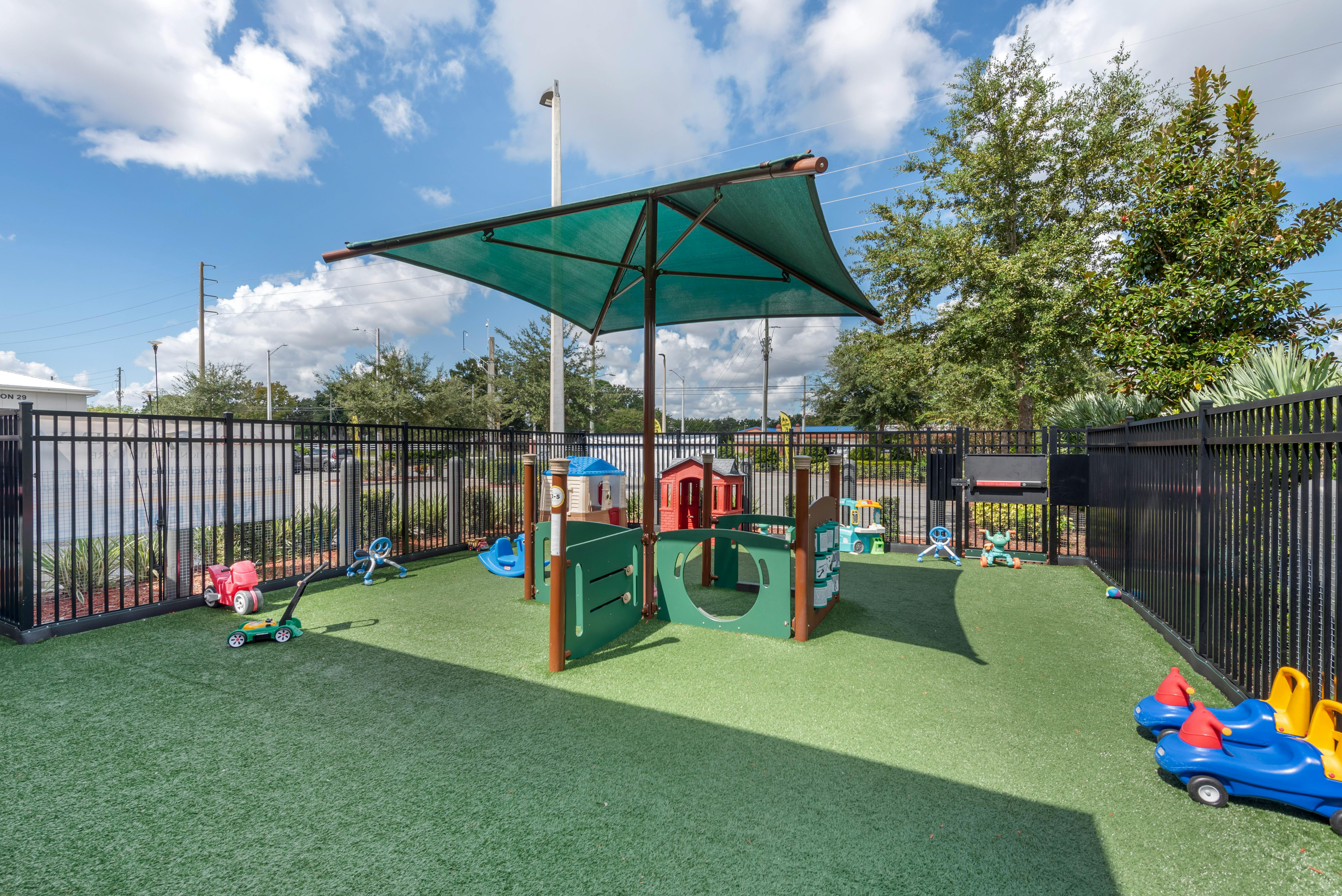 Outdoor playground with green turf, play structures, and a large shade canopy