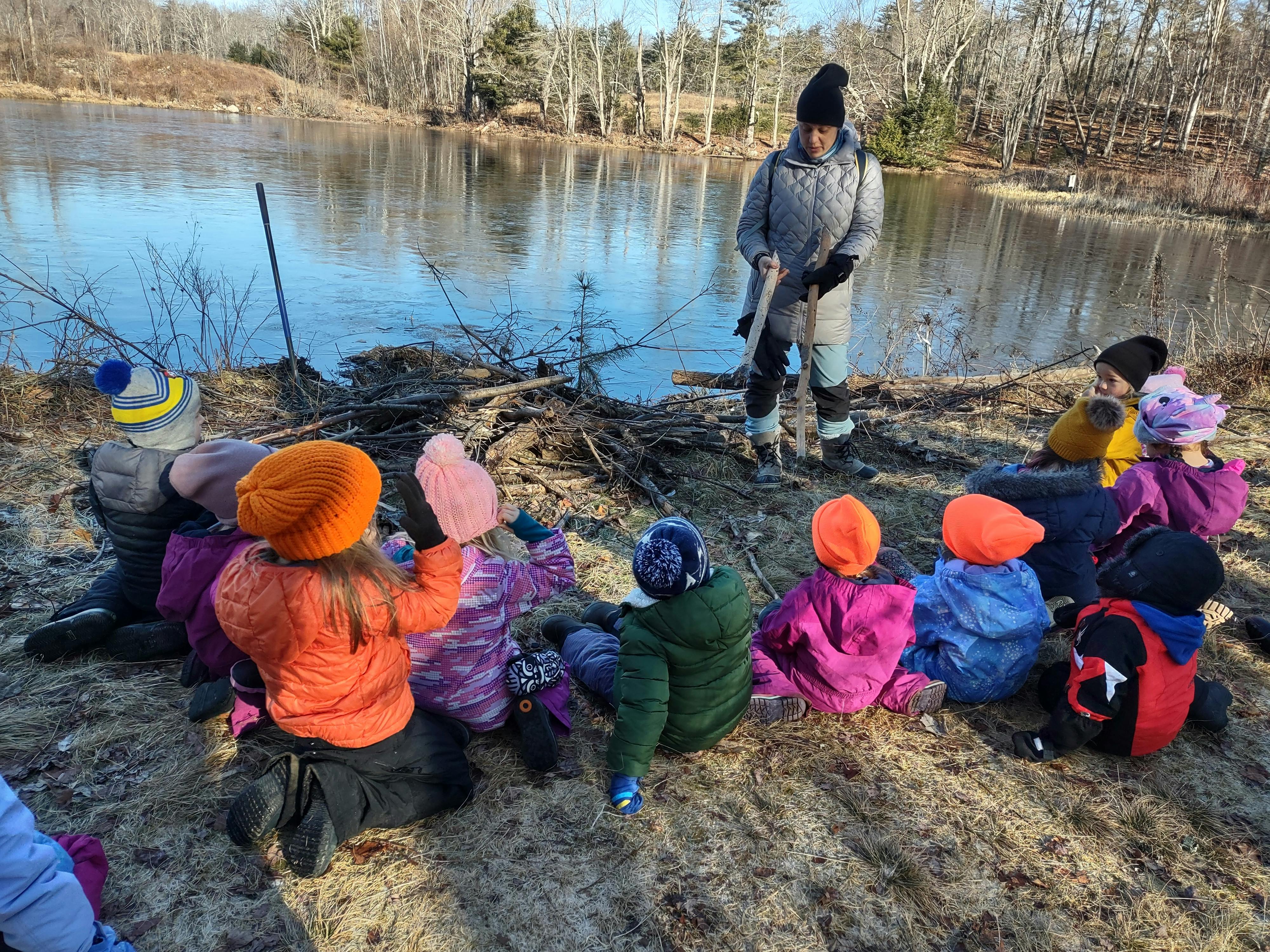 Person standing by a lake with children sitting on the ground around them