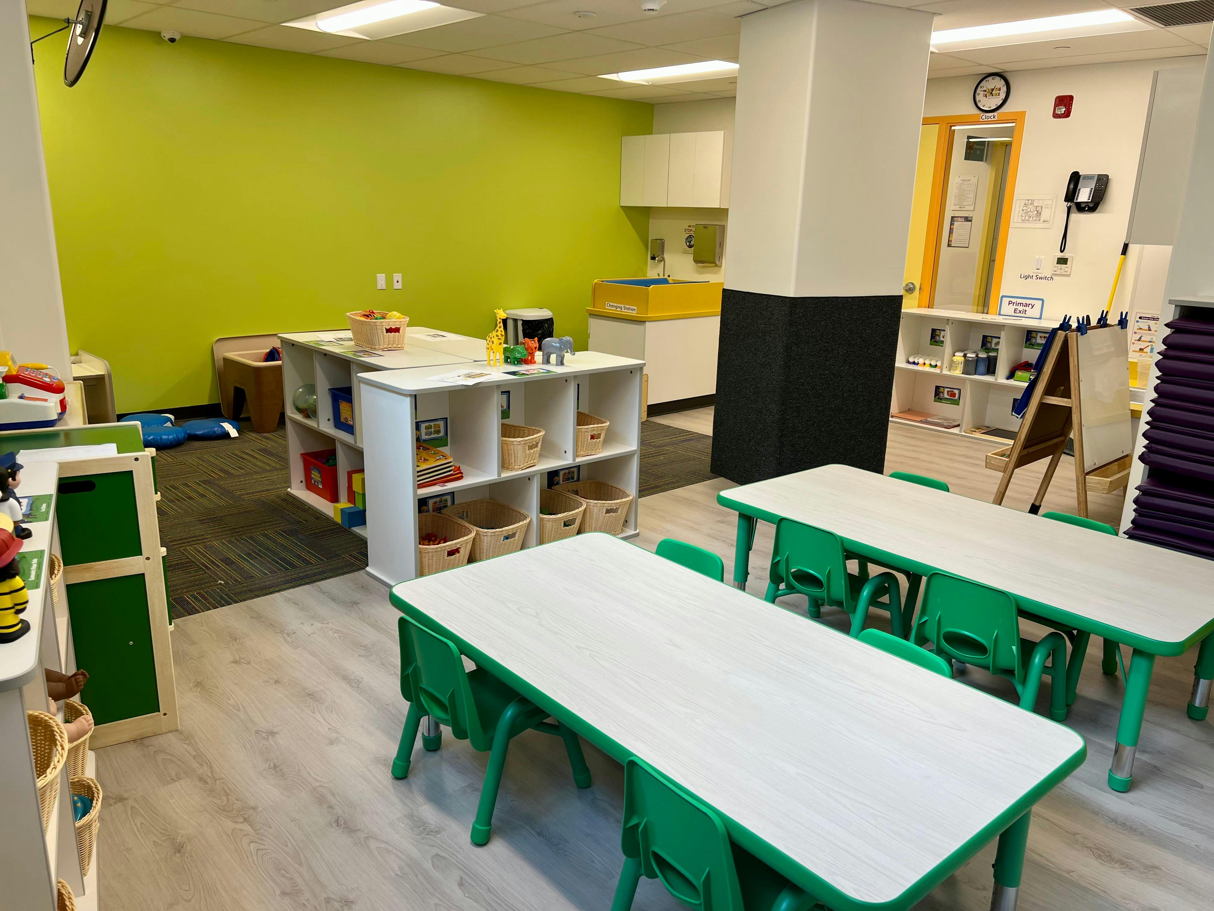 Classroom with small tables and chairs, shelves with baskets, and a green wall