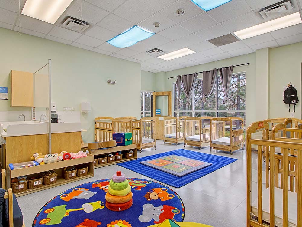 A brightly lit nursery with multiple wooden cribs, changing tables, and colorful rugs