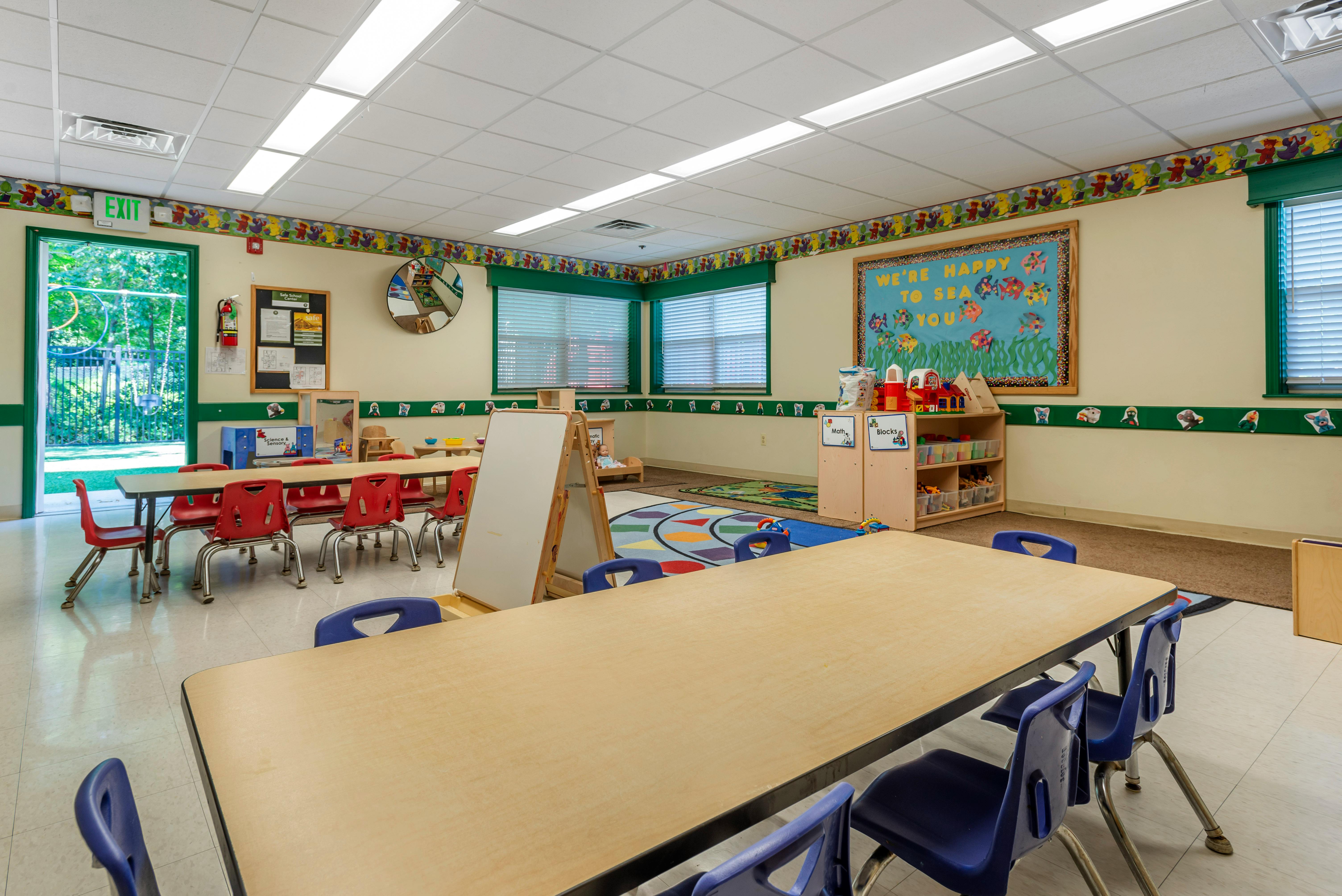 Classroom with child-sized tables and chairs, a whiteboard, and an exit door