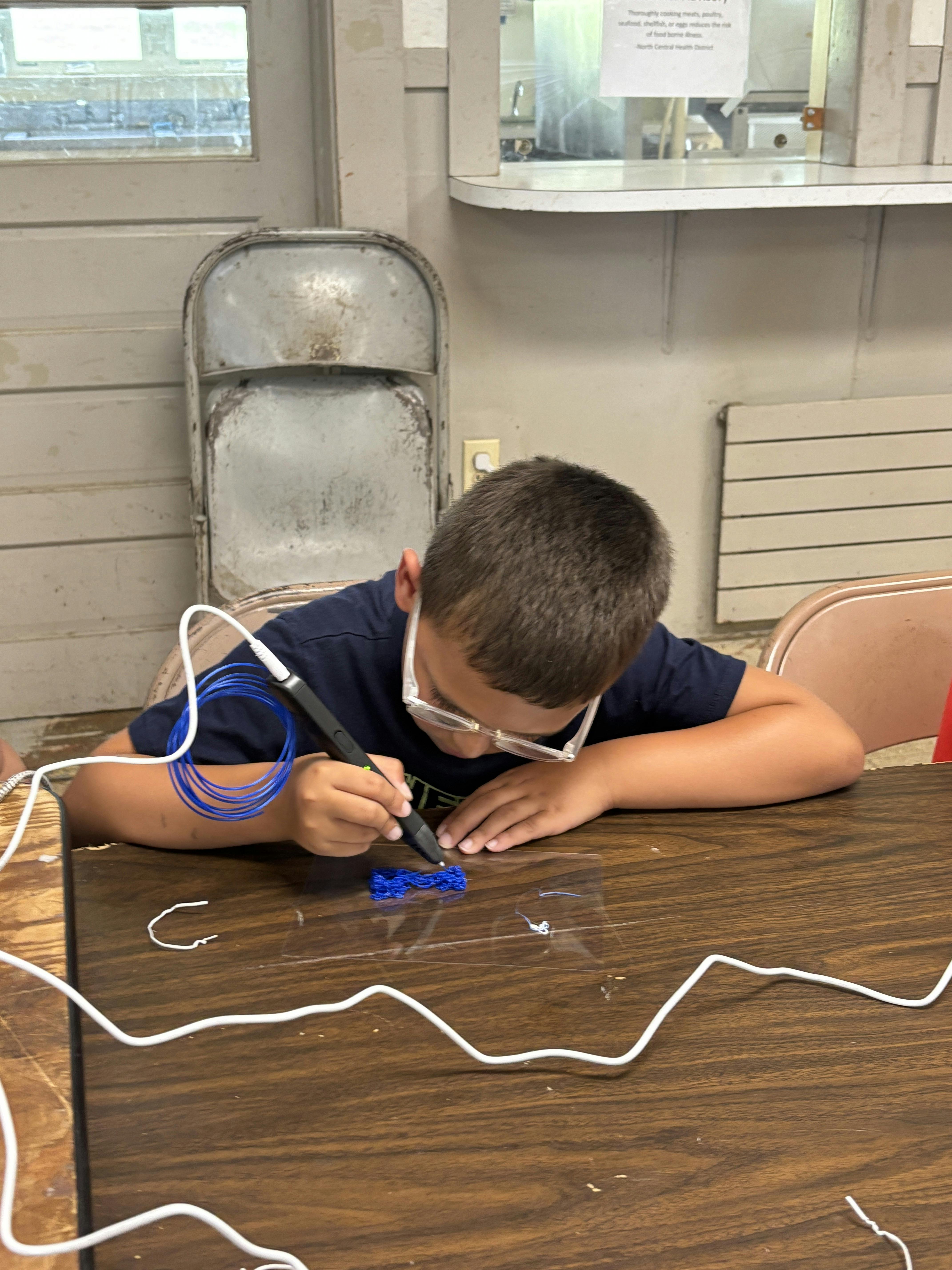 Child wearing glasses using a 3D printing pen to create a blue object