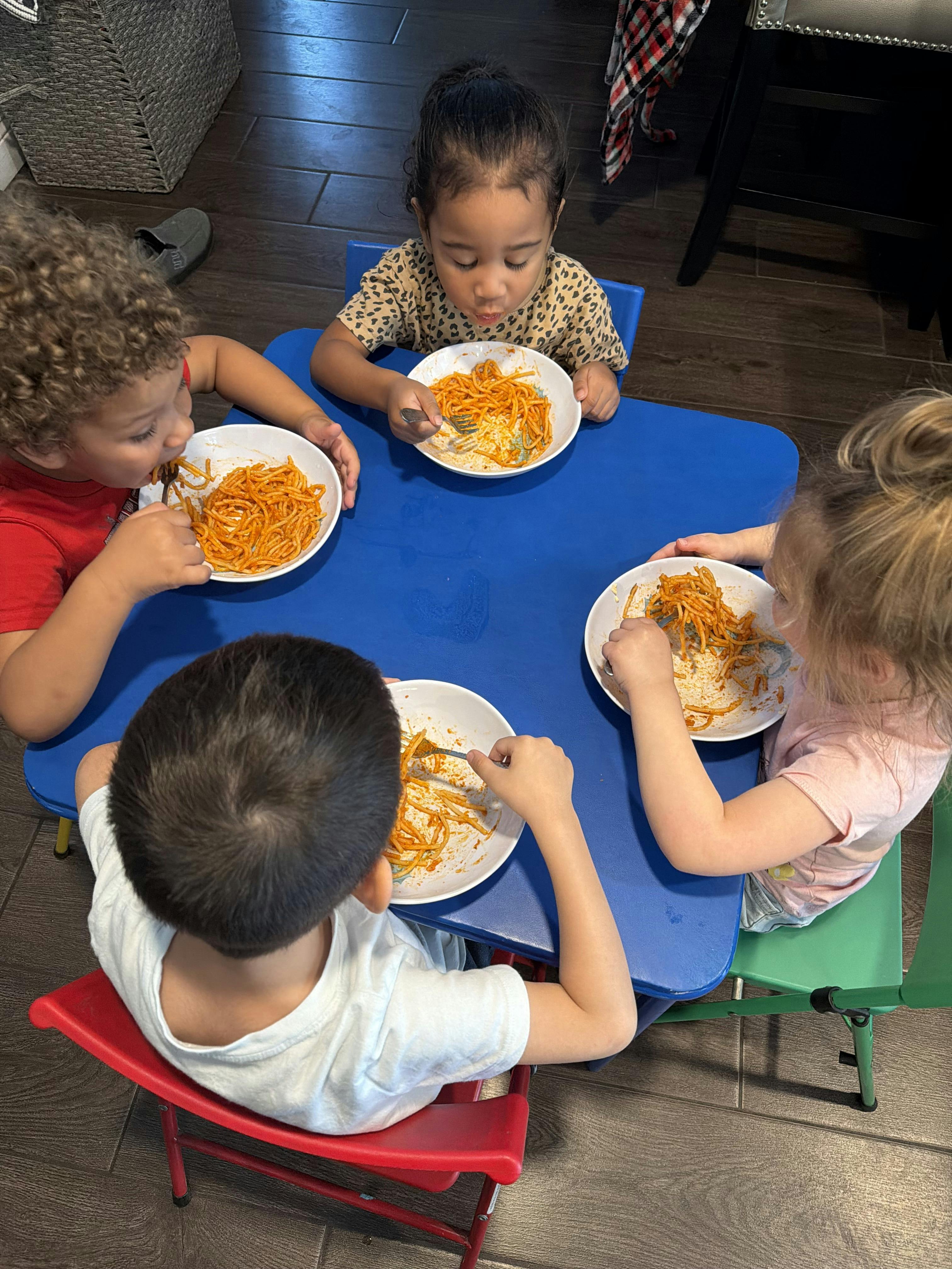 Four children eating spaghetti from white bowls at a blue table