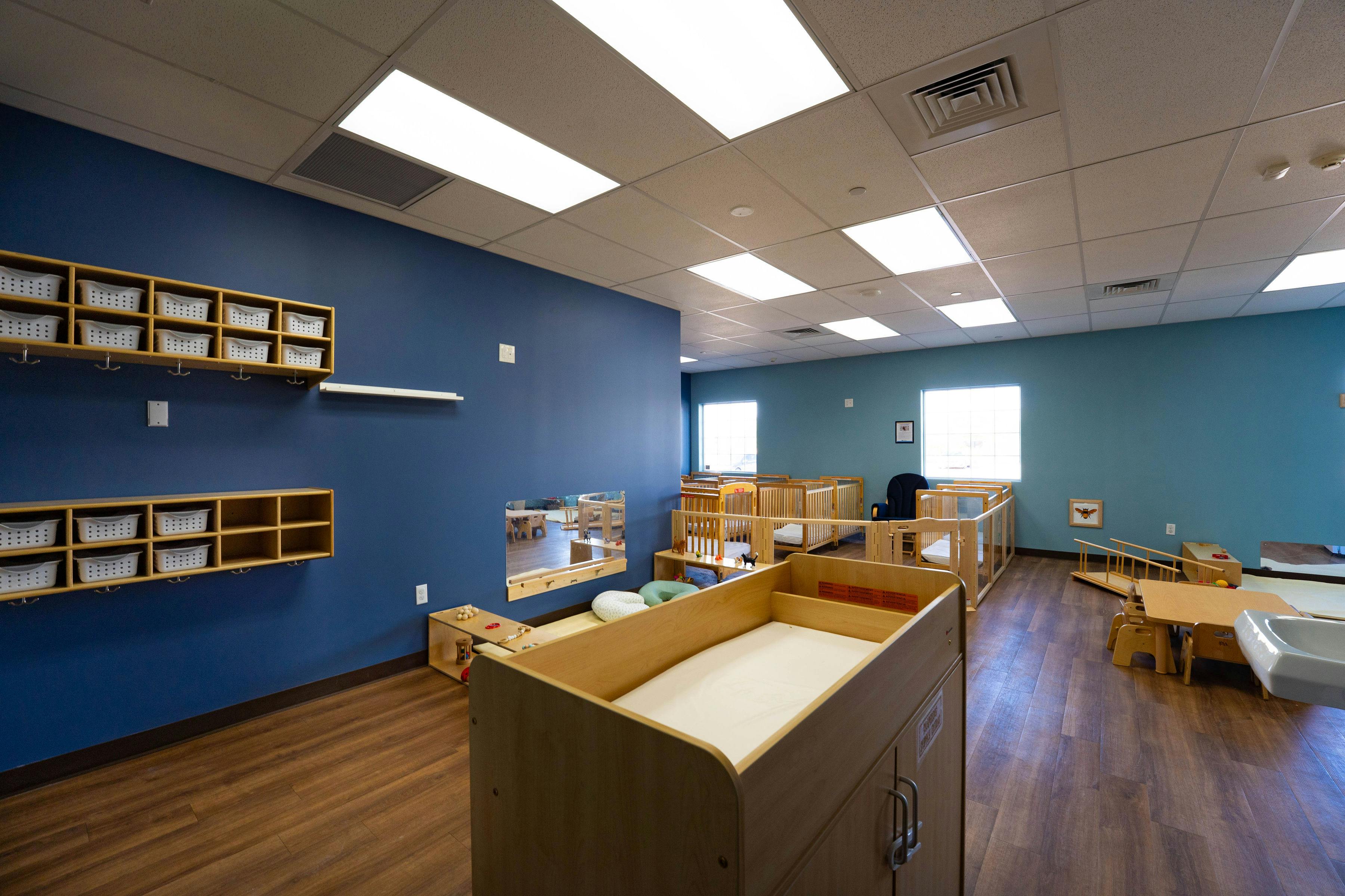 A daycare room with blue and teal walls, cribs, changing table, and storage cubbies
